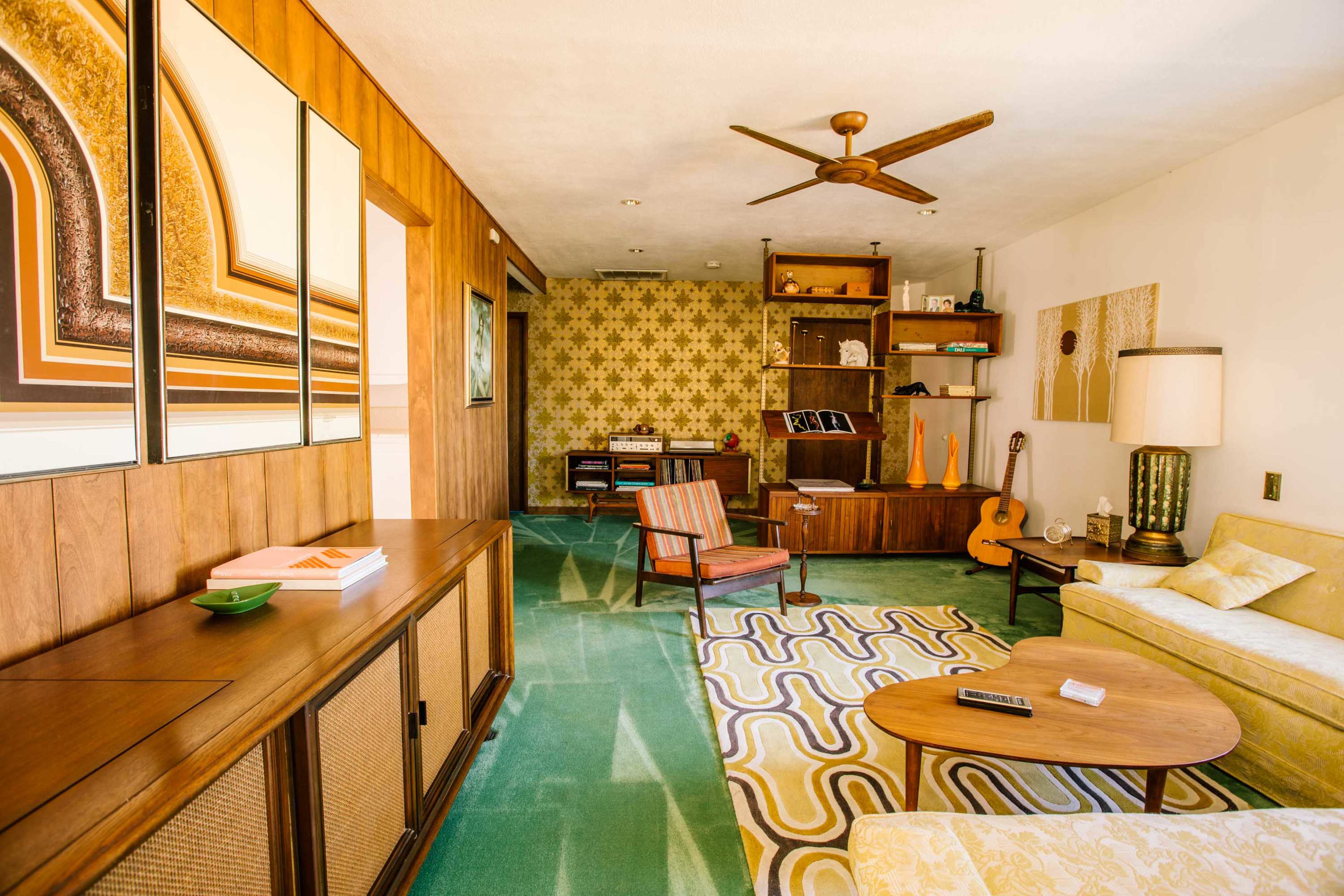 The image shows a mid-century modern living room featuring wooden paneling, a green carpet, and various furnishings including a guitar, a record player, and a patterned area rug.