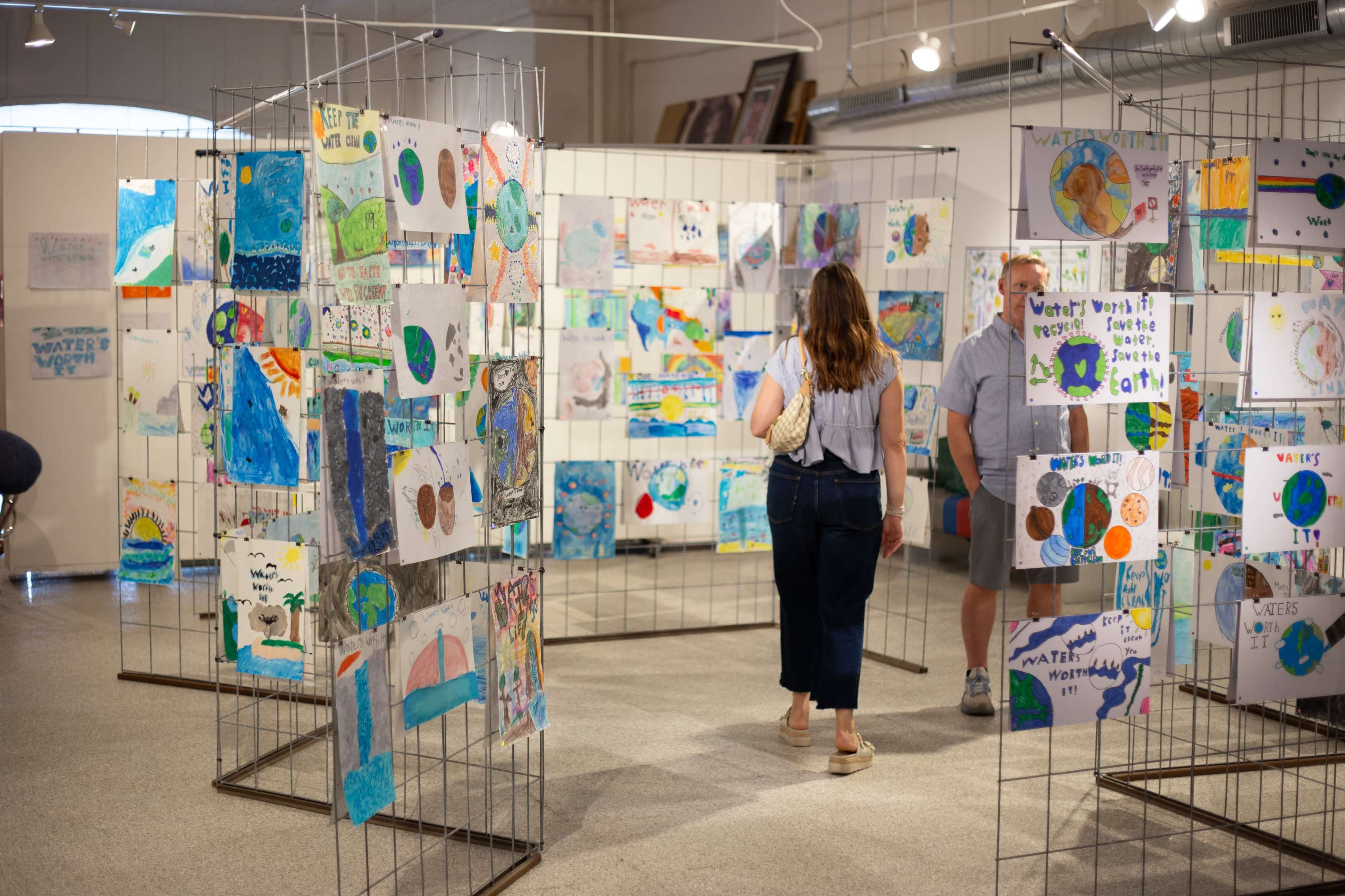 People are walking through an art gallery displaying colorful children's artwork on partitioned walls.