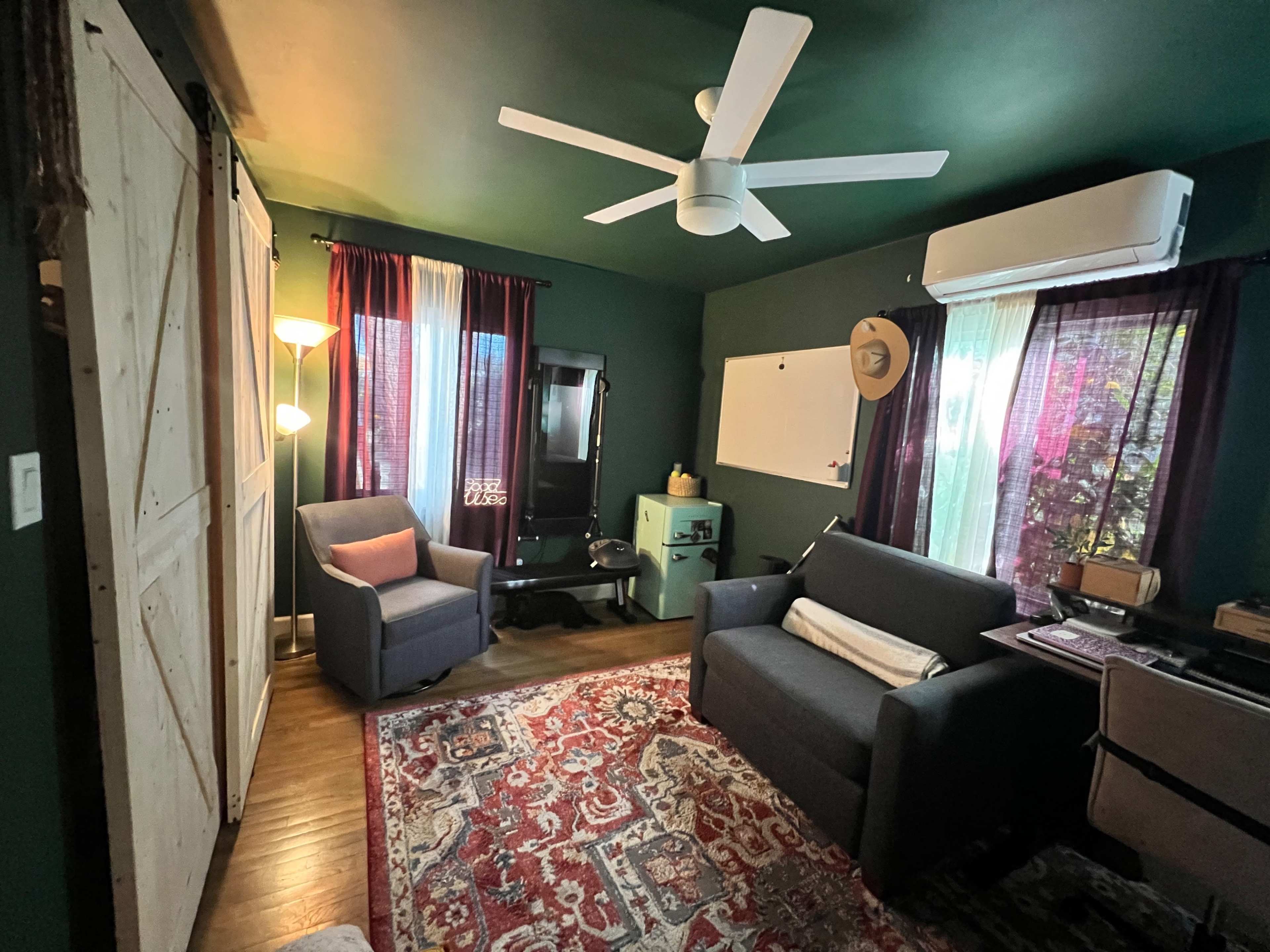 A cozy room with a green-painted wall, a ceiling fan, a dark rug, two armchairs, a small table, and large windows with curtains.