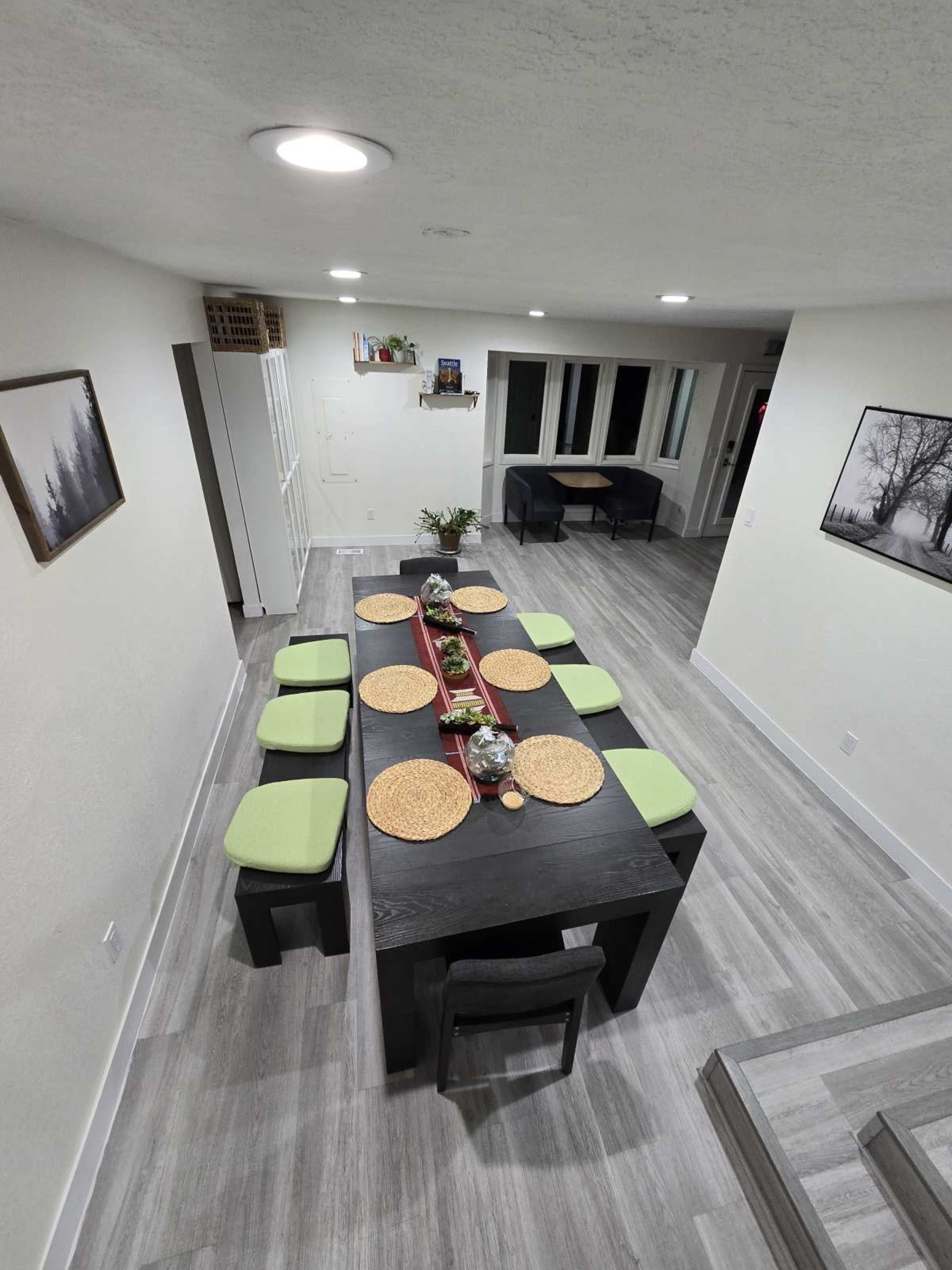 The image shows a modern dining area with a long black table set for eight, featuring circular placemats and green cushions, surrounded by light-colored walls and large windows.