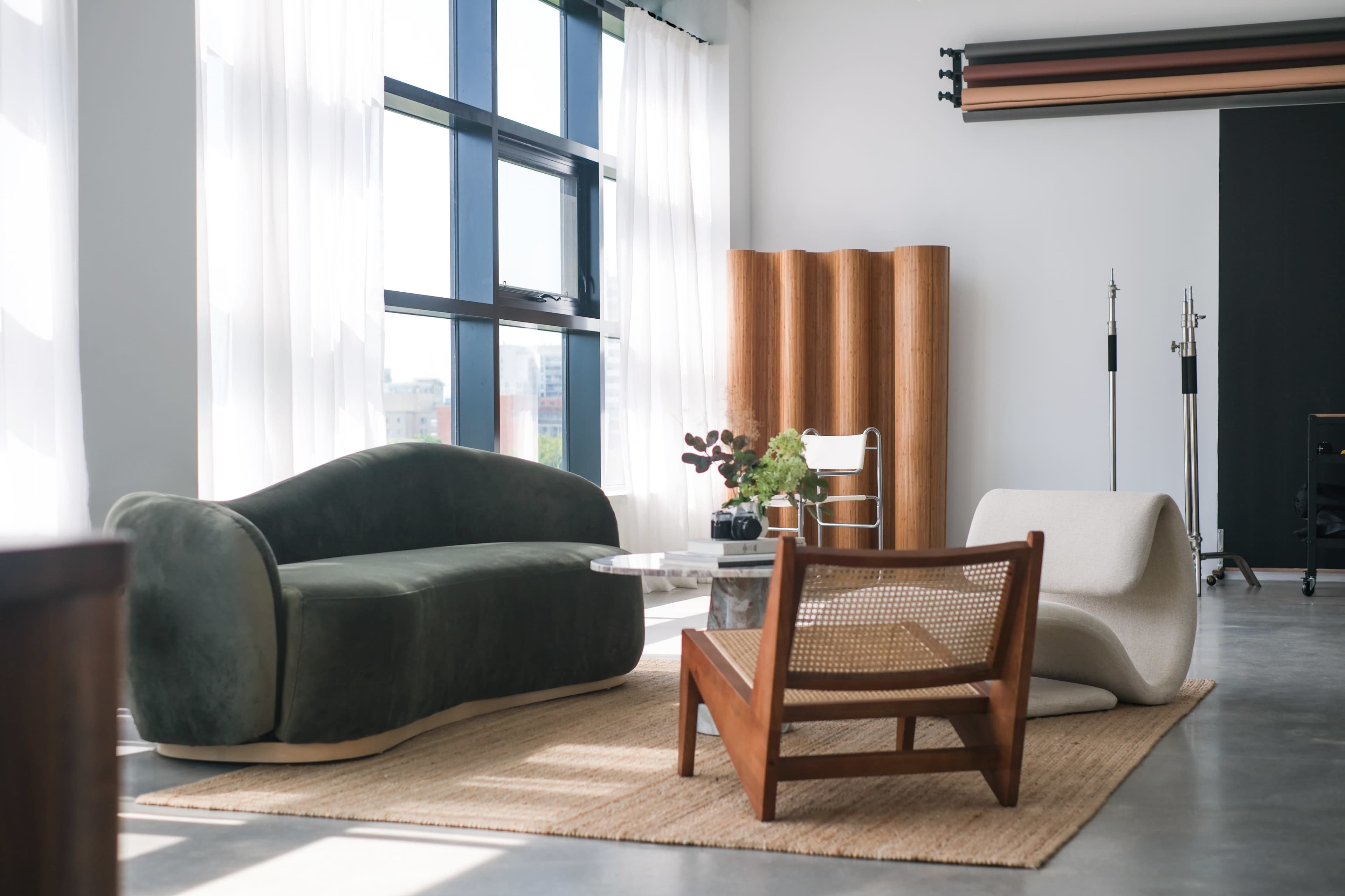 The image shows a modern, well-lit living space featuring a green sofa, a white lounge chair, and a wooden accent chair on a textured rug.