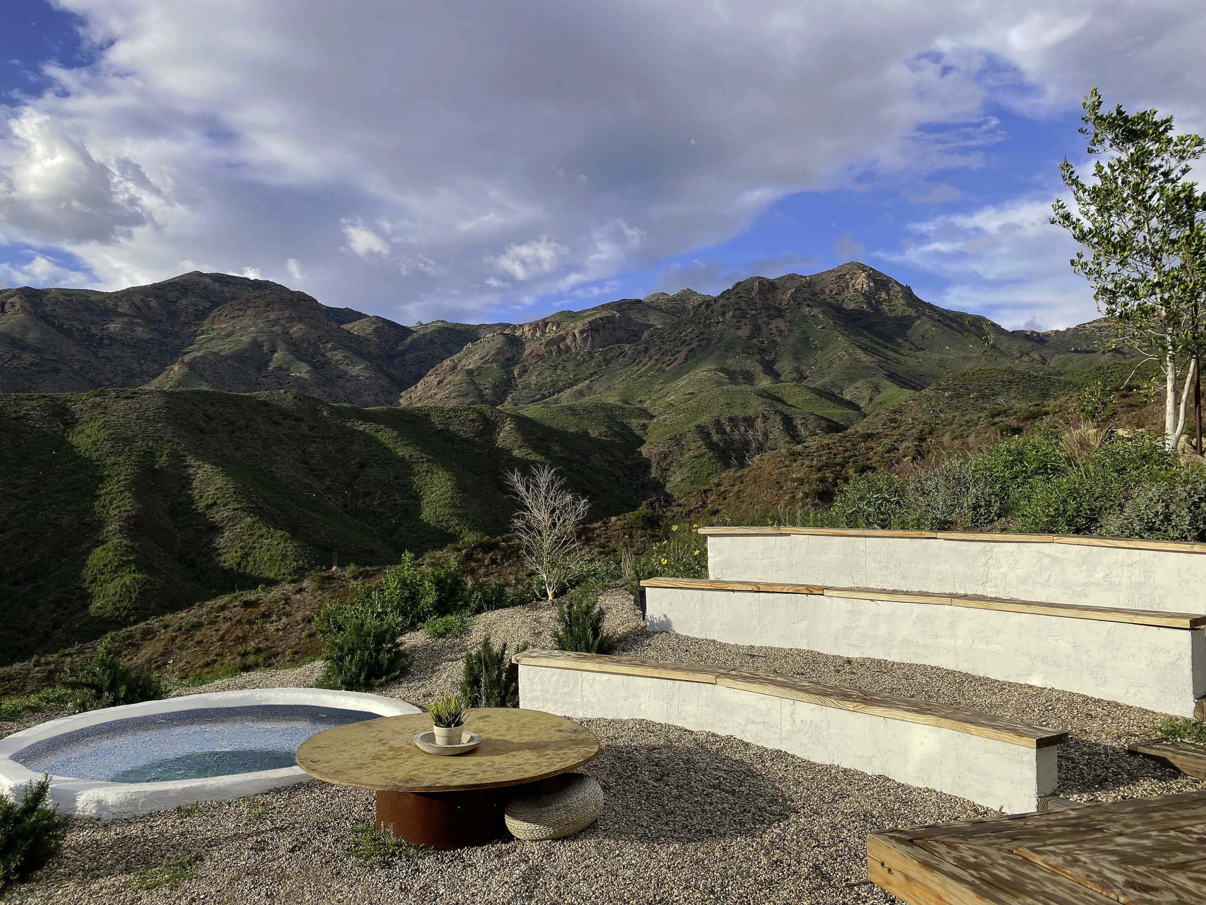 A terraced seating area made of stone and wood overlooks a mountain landscape with sparse vegetation and a small circular pool.