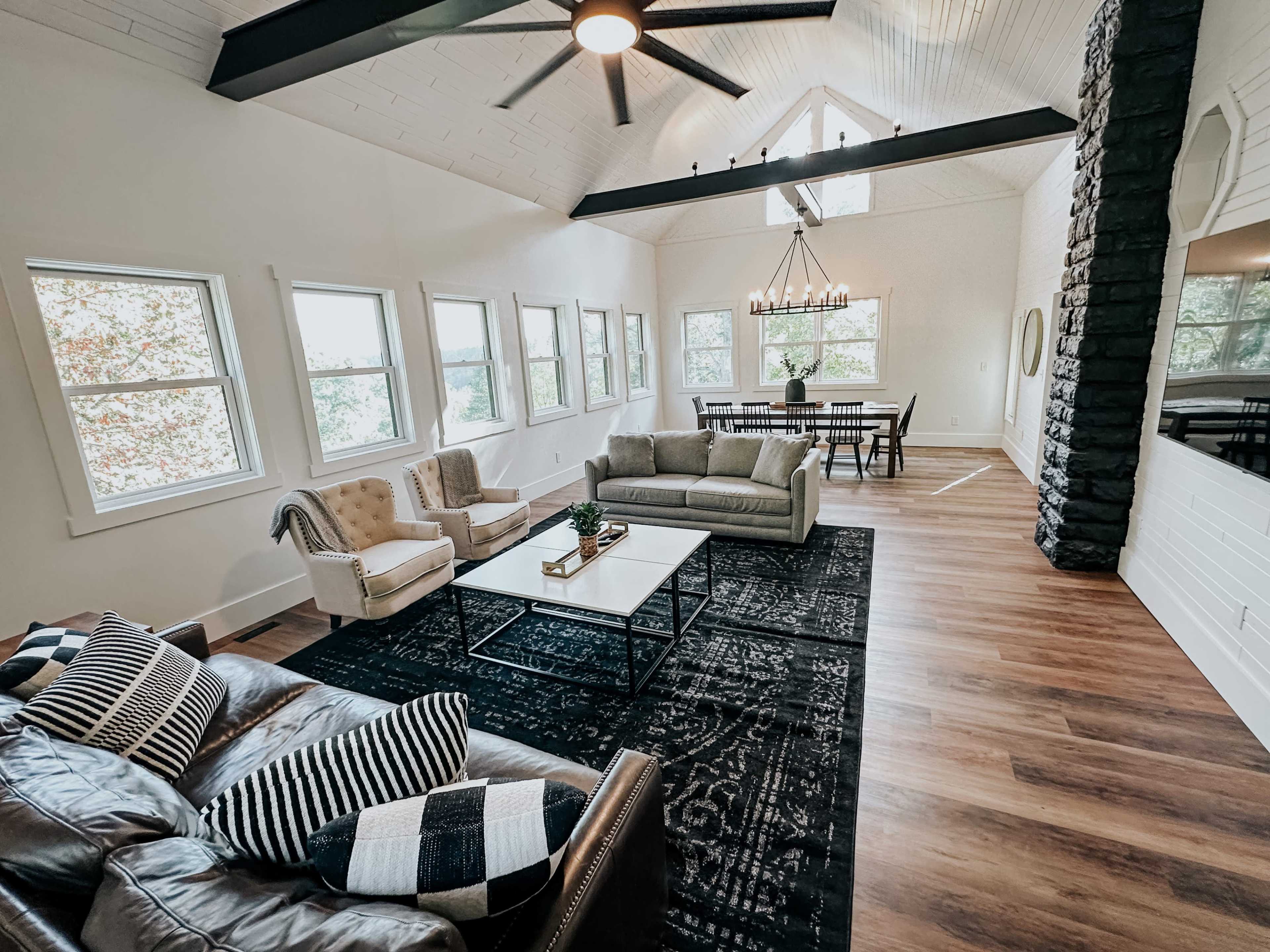 A spacious living room with large windows, modern furniture, a central coffee table, and a dining area in the background.