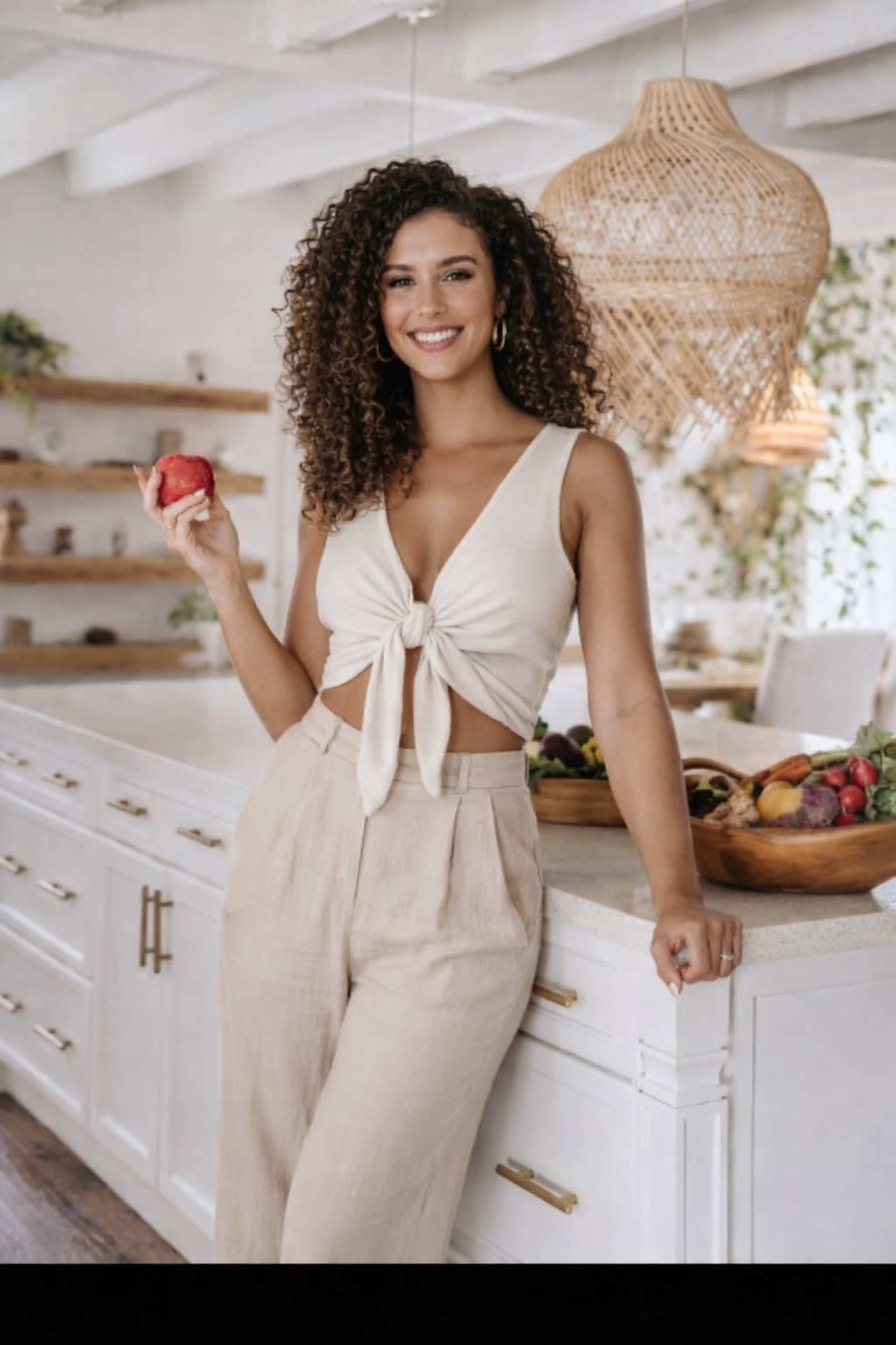 A woman stands in a bright kitchen, holding an apple and smiling, with fruits displayed in a wooden bowl nearby.