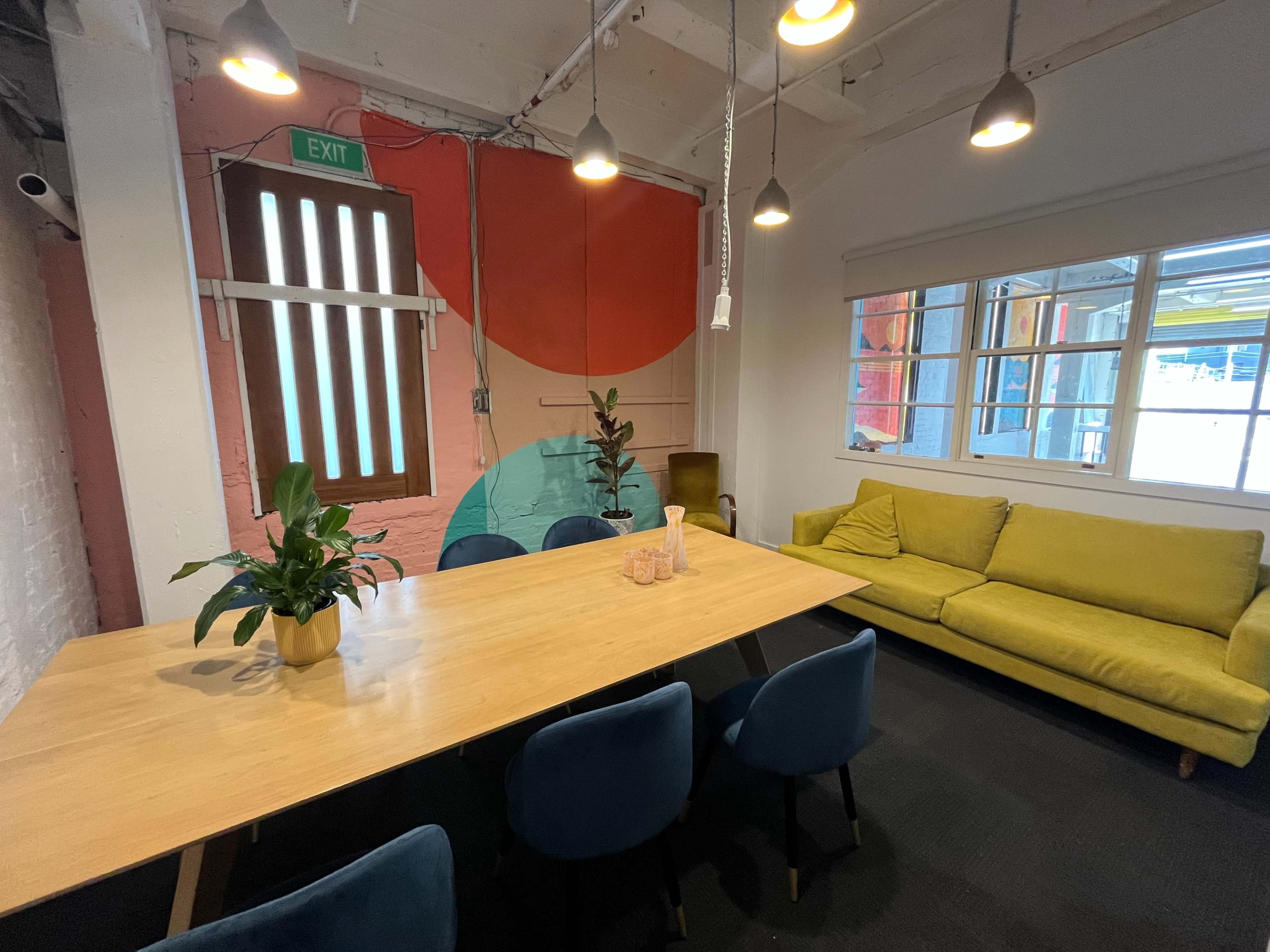 A modern meeting room features a large wooden table, blue chairs, a yellow couch, and colorful wall art with plants decor.