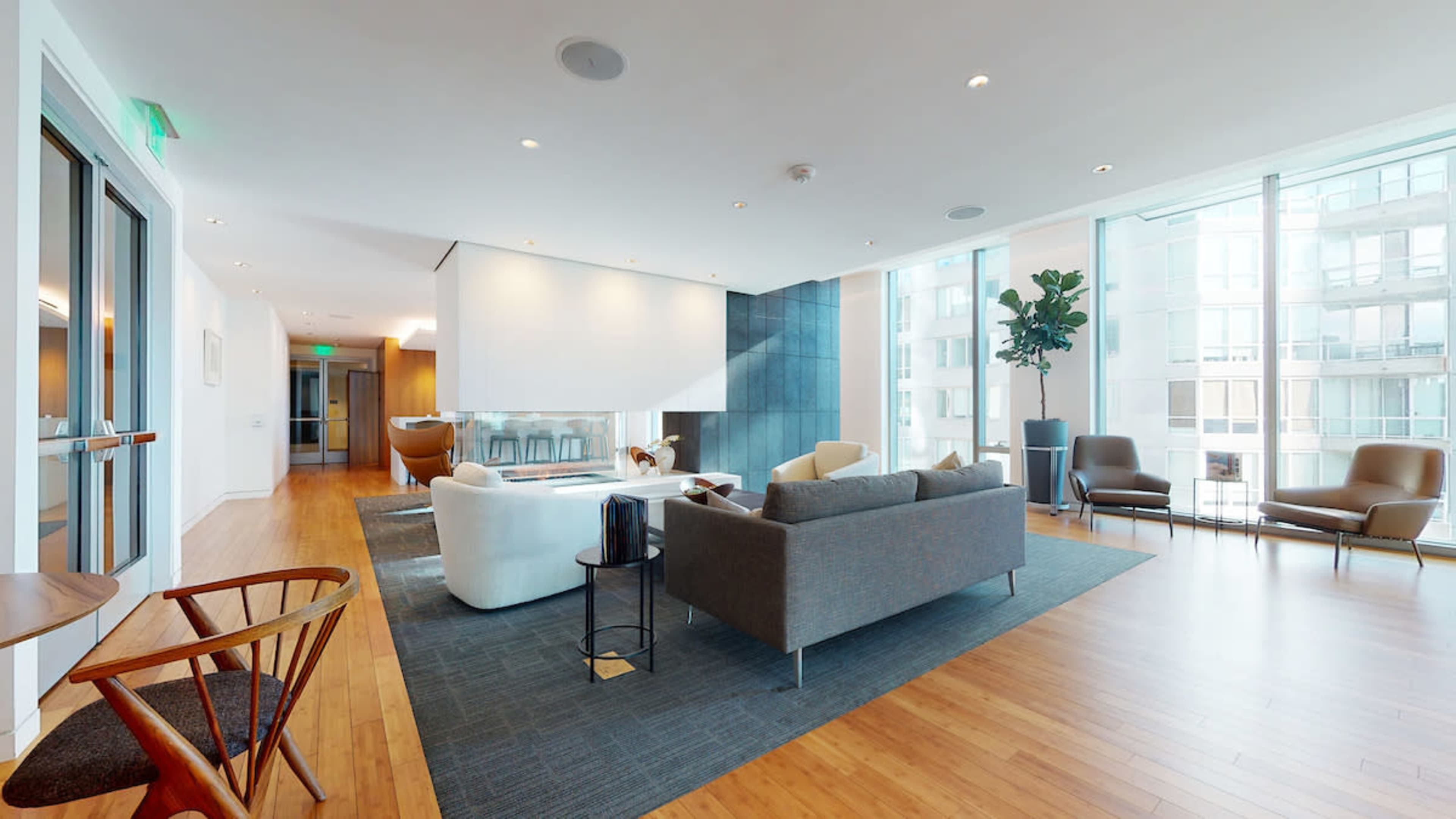 A modern, brightly lit lounge area with large windows, featuring a mix of comfortable seating arrangements and a decorative plant.