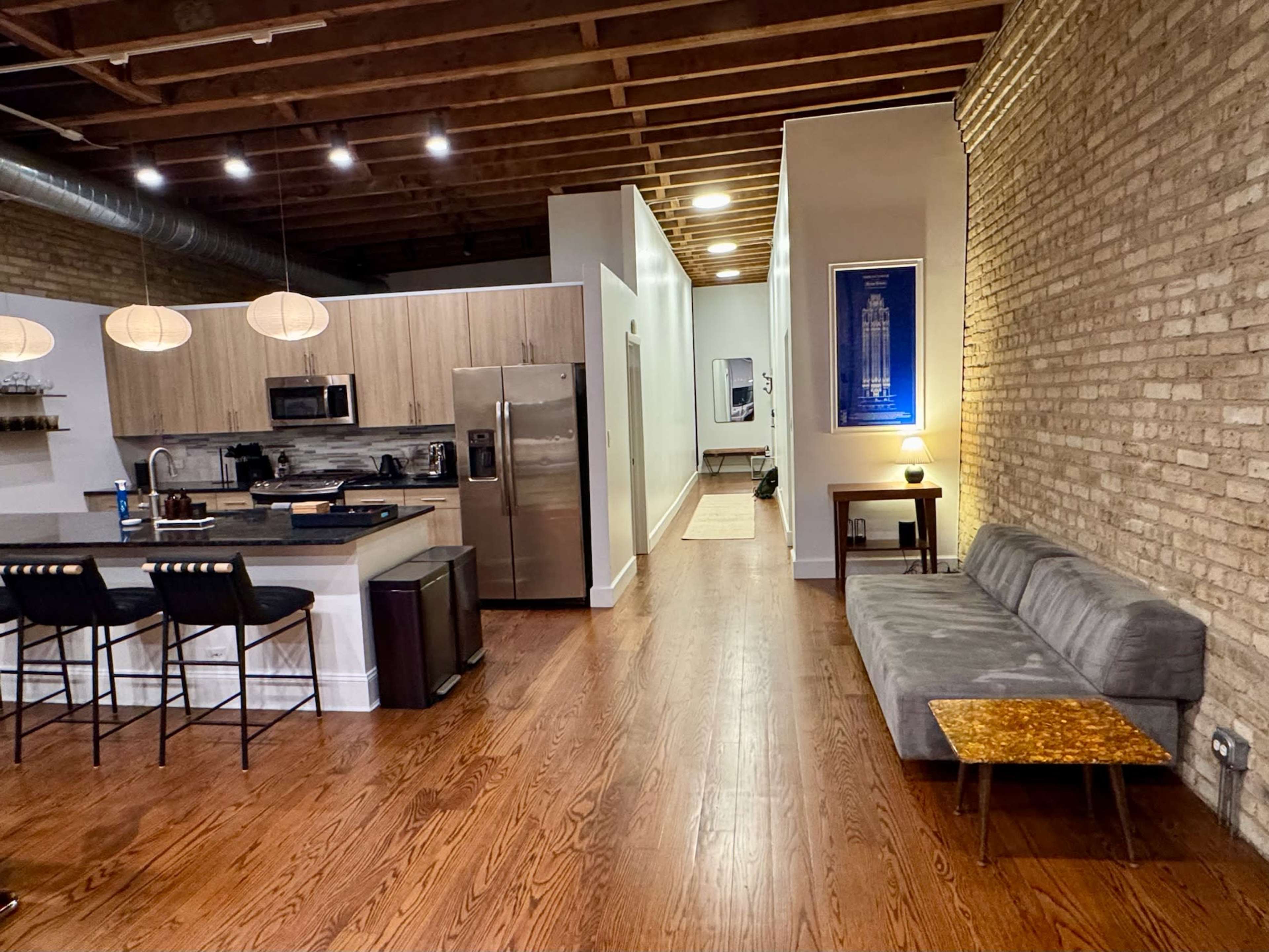 The image shows a modern open-concept living space featuring a kitchen with stainless steel appliances and a cozy seating area with a gray sofa and wooden accents.