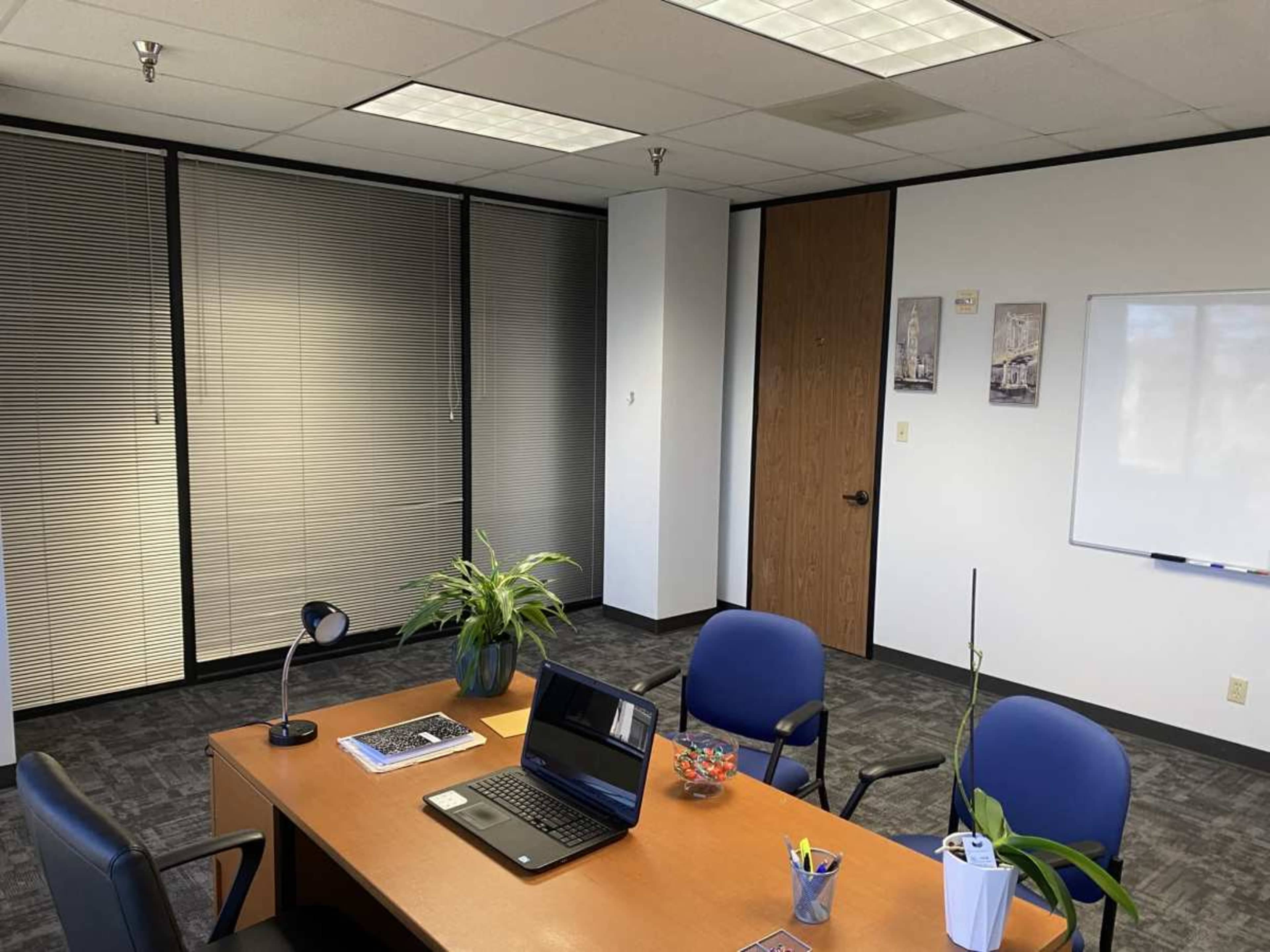 The image shows a modern office space with a wooden desk, two blue chairs, a laptop, a lamp, a plant, and a whiteboard on the wall.