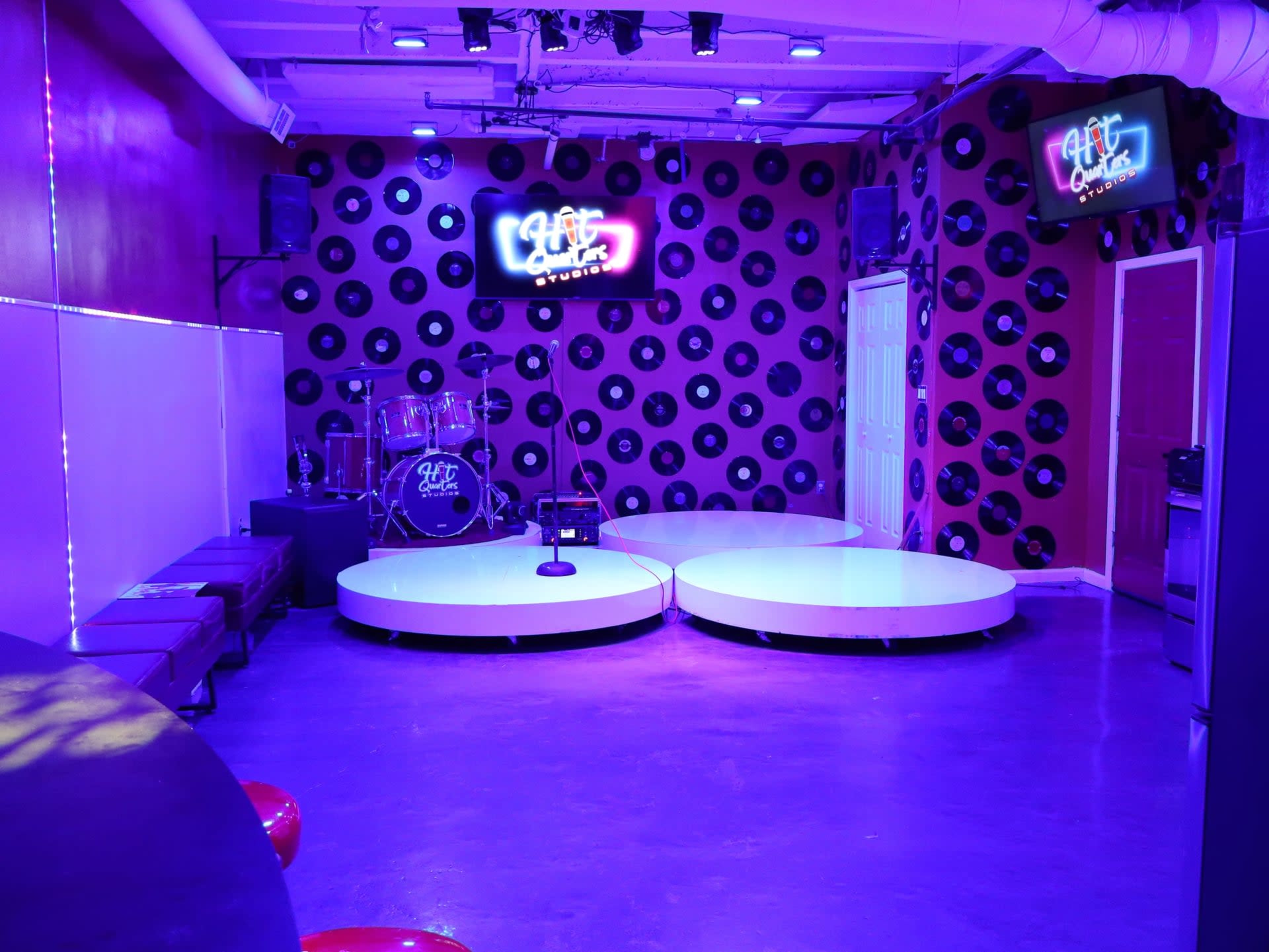The room features a stage with circular platforms, a drum set, and walls adorned with vinyl records, illuminated by purple lighting.