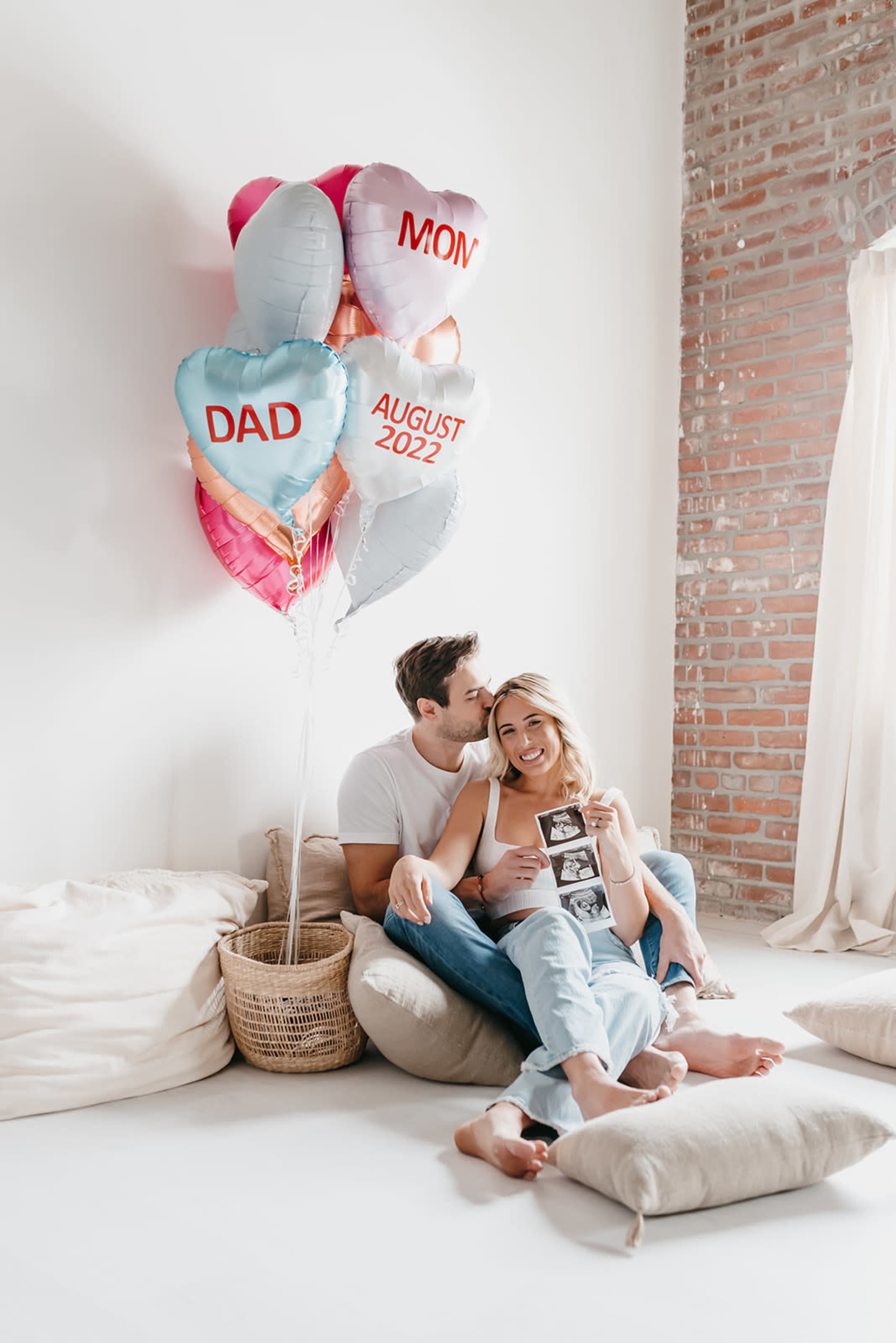 A couple sits together on cushions, surrounded by heart-shaped balloons labeled "MON," "DAD," and "AUGUST 2022," while holding an ultrasound photo.