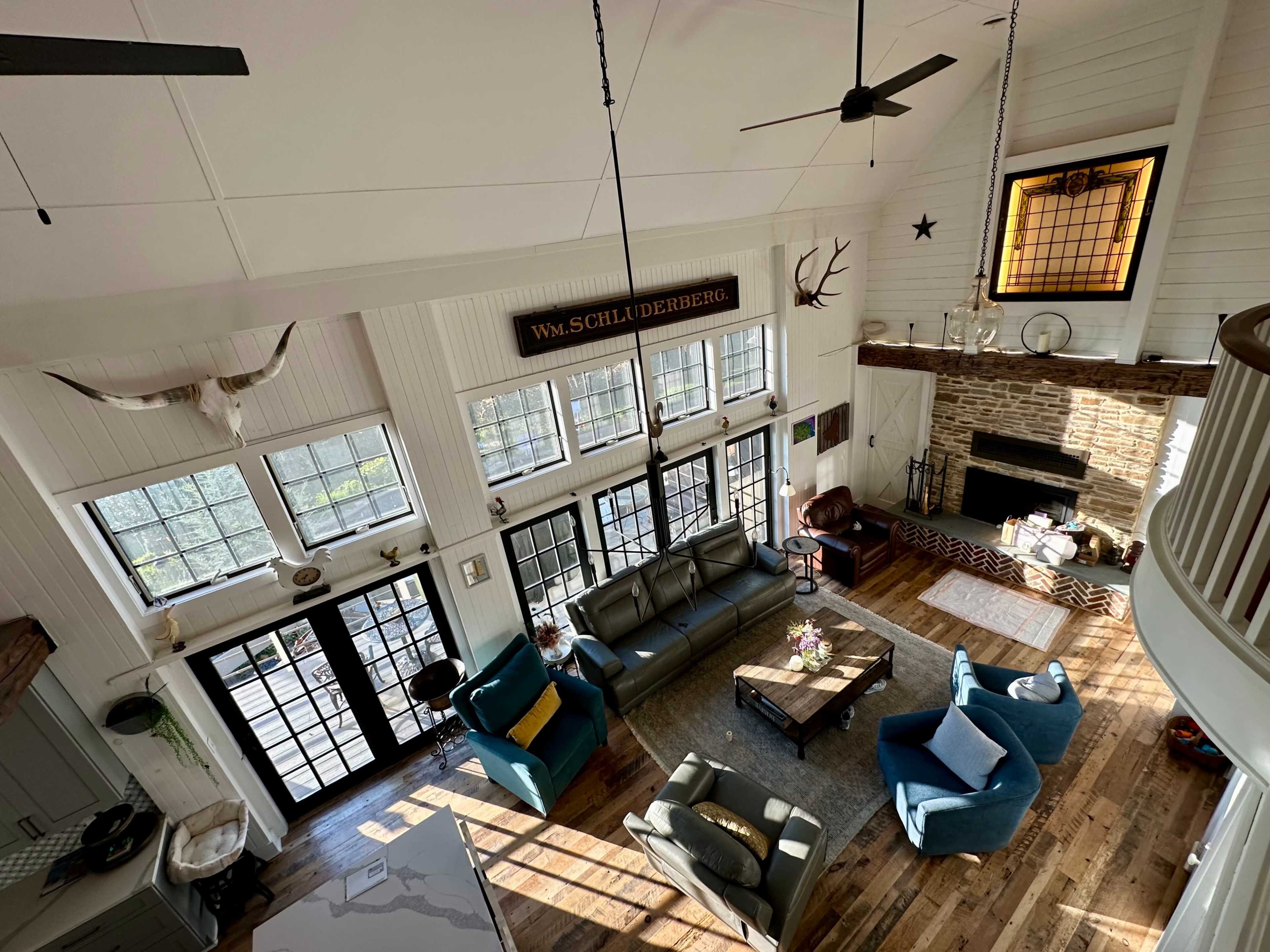 The image shows a spacious living room with large windows, a stone fireplace, and a mix of modern and rustic furniture.