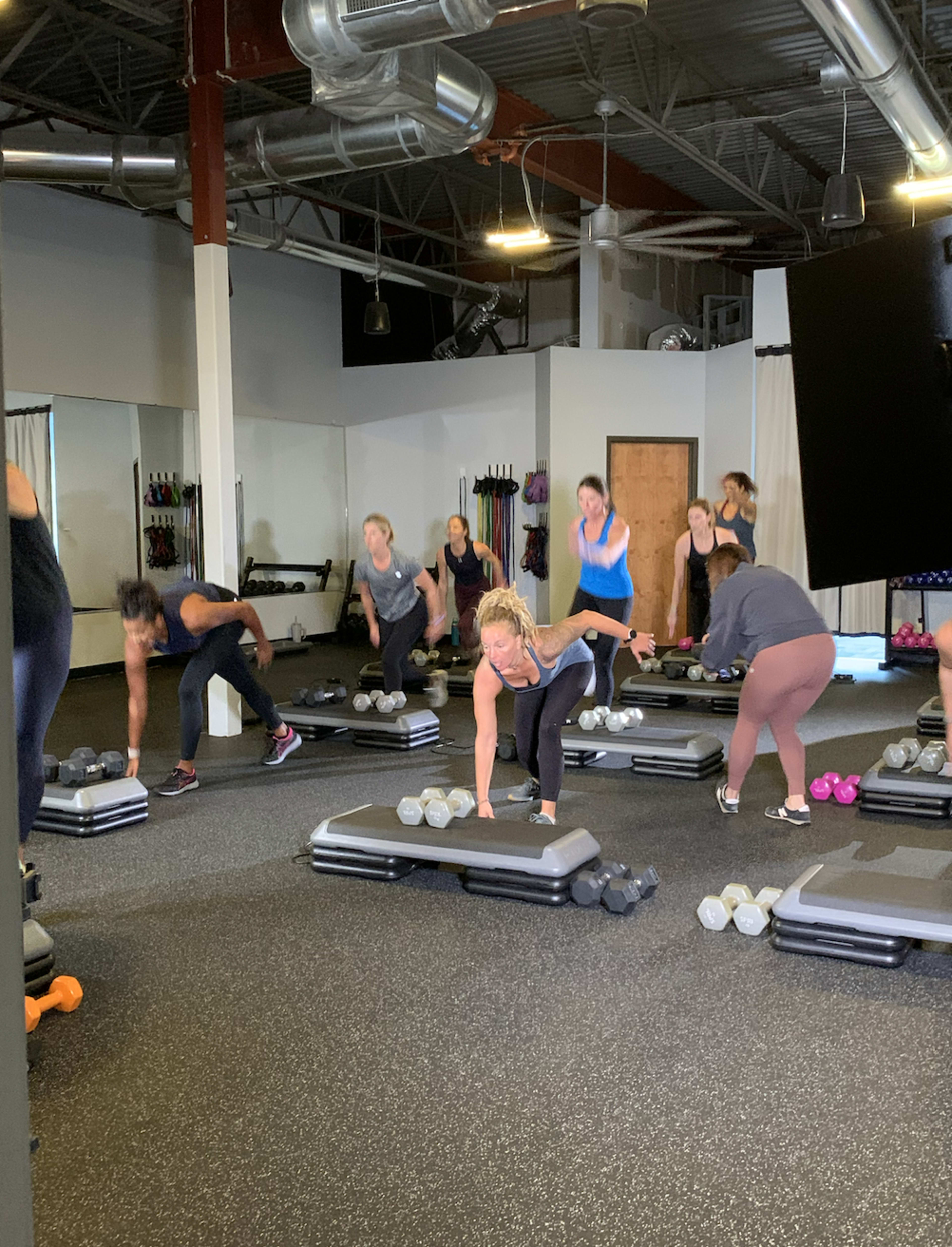 A group of people exercises in a fitness studio while using weights and step platforms.