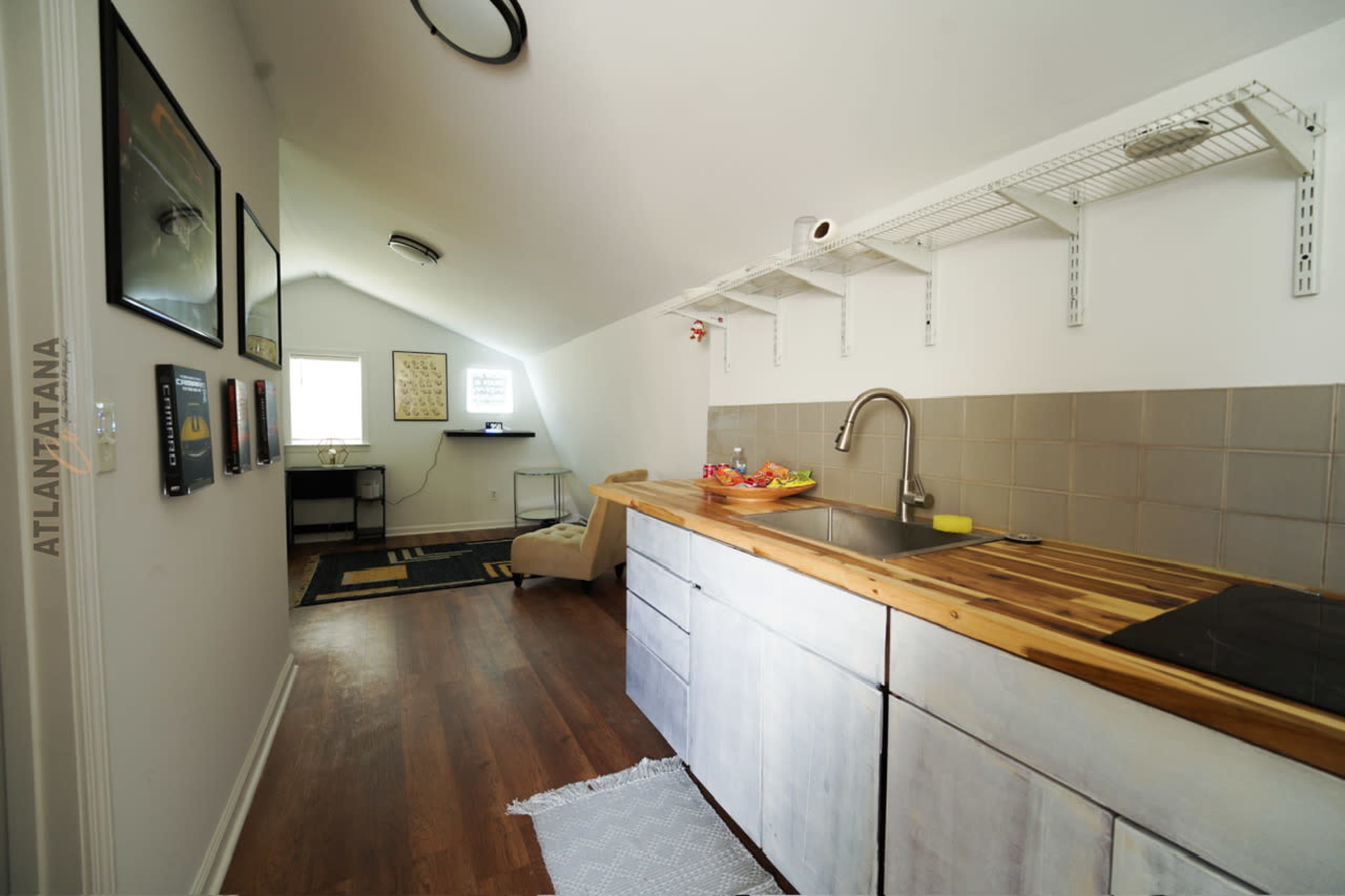 The image shows a modern kitchen area with wooden countertops, gray cabinets, and a small dining space featuring a chair and a wall-mounted shelf.