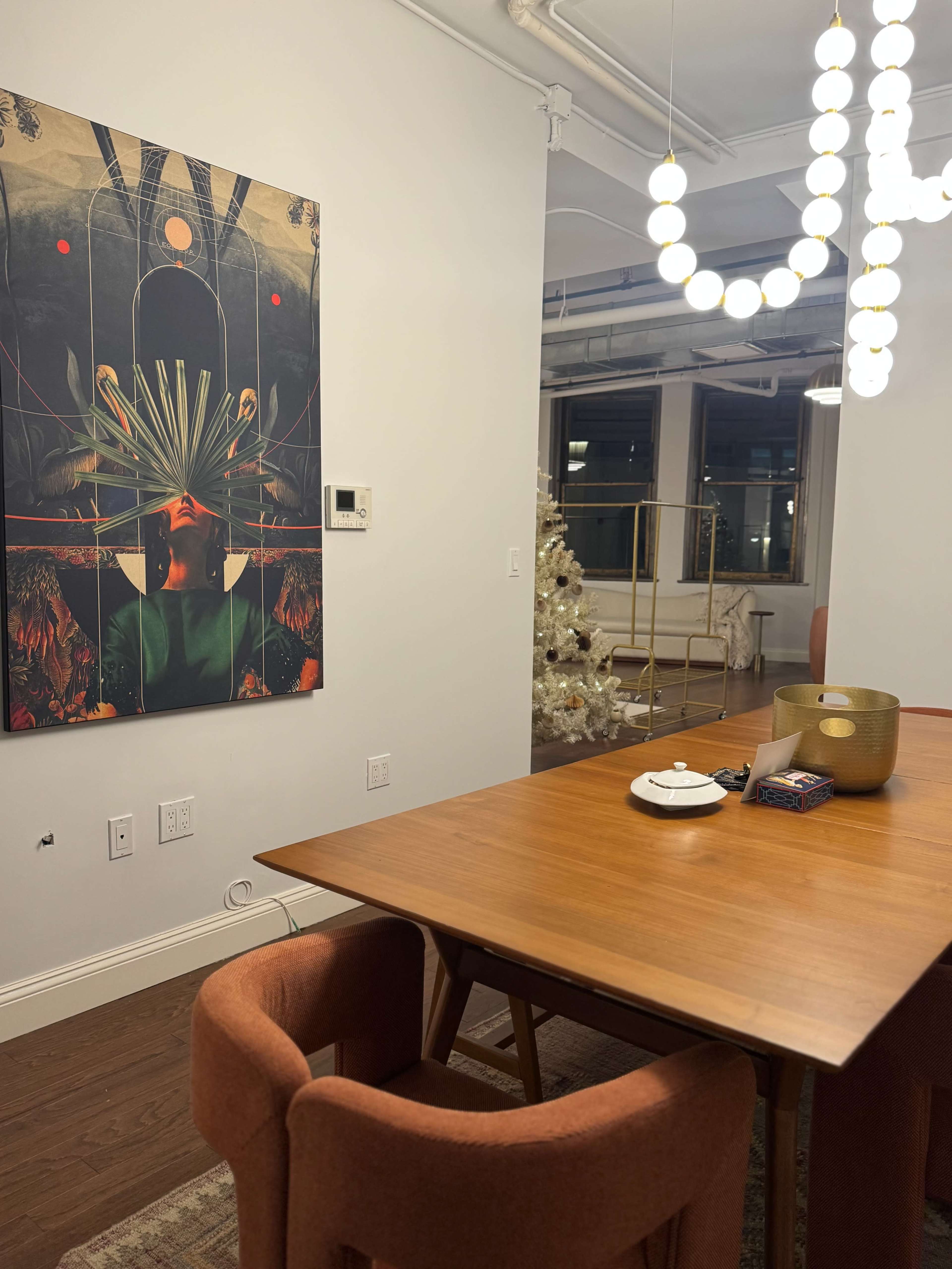 A modern dining area features a wooden table surrounded by upholstered chairs, with a large piece of art on the wall and a decorative Christmas tree in the background.