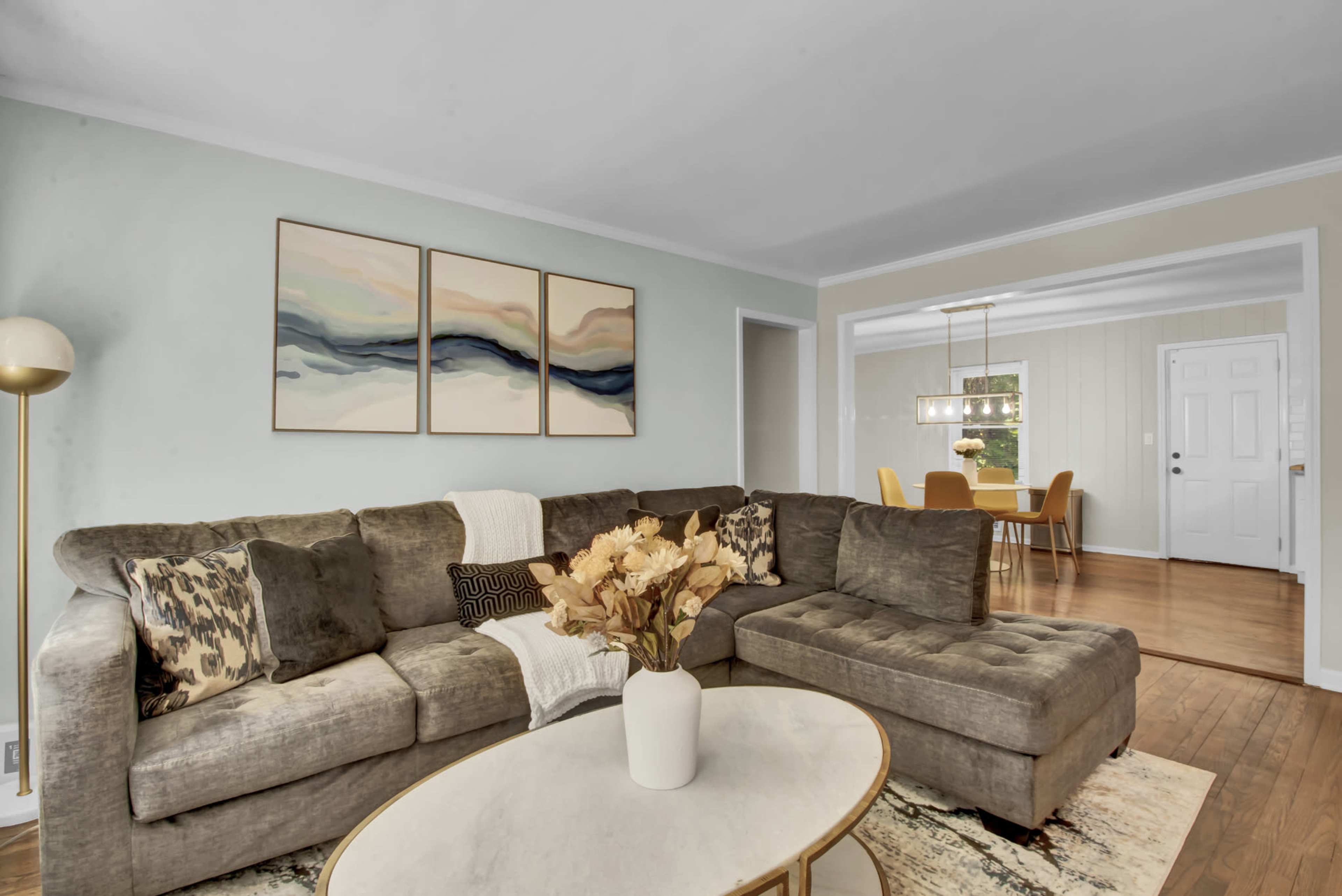 The image shows a living room with a sectional sofa, a round coffee table with a vase of flowers, and three abstract paintings on the wall opposite an adjacent dining area.