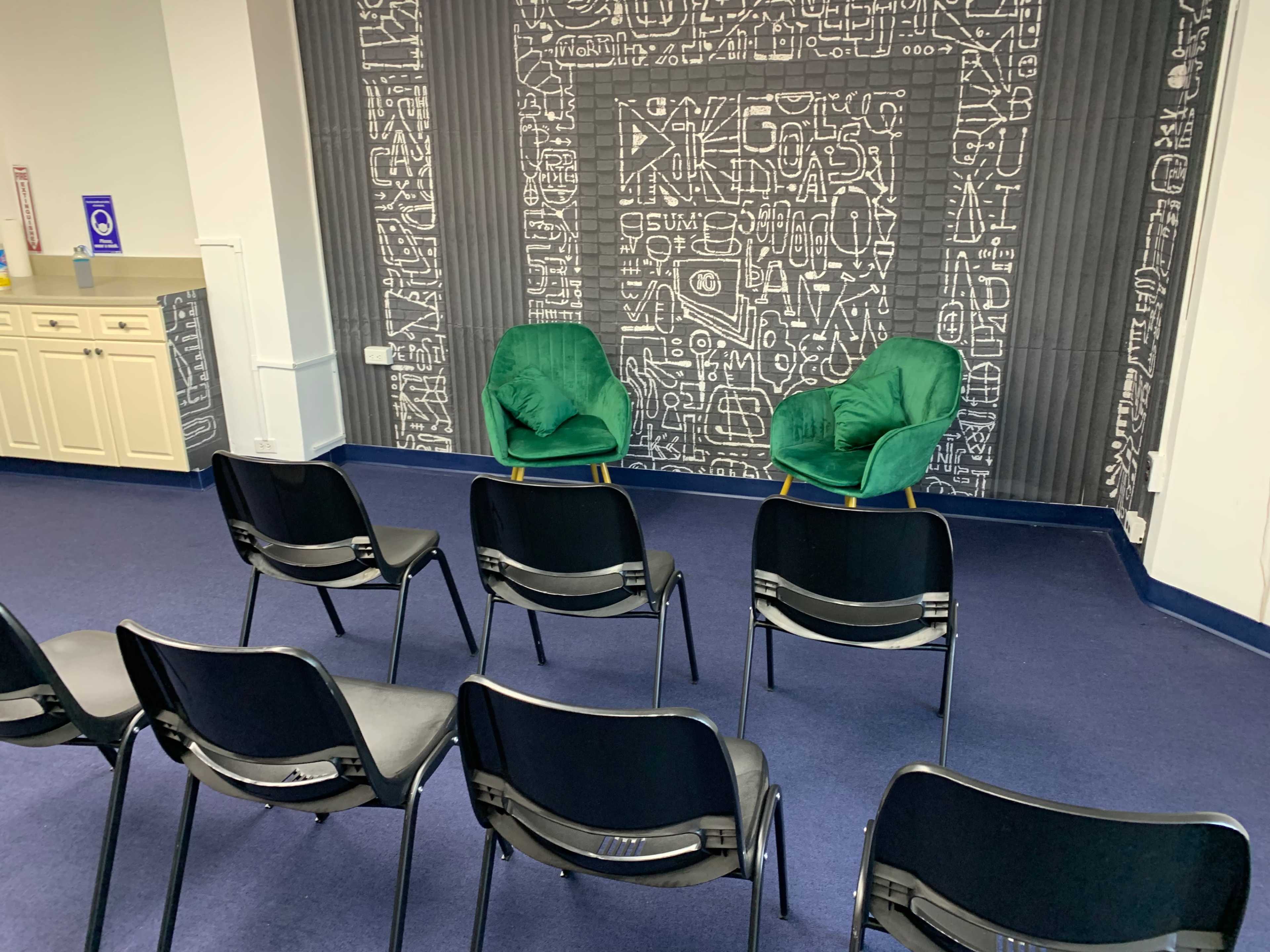 Two green chairs are positioned in front of a patterned wall, with several black chairs arranged in rows facing them.