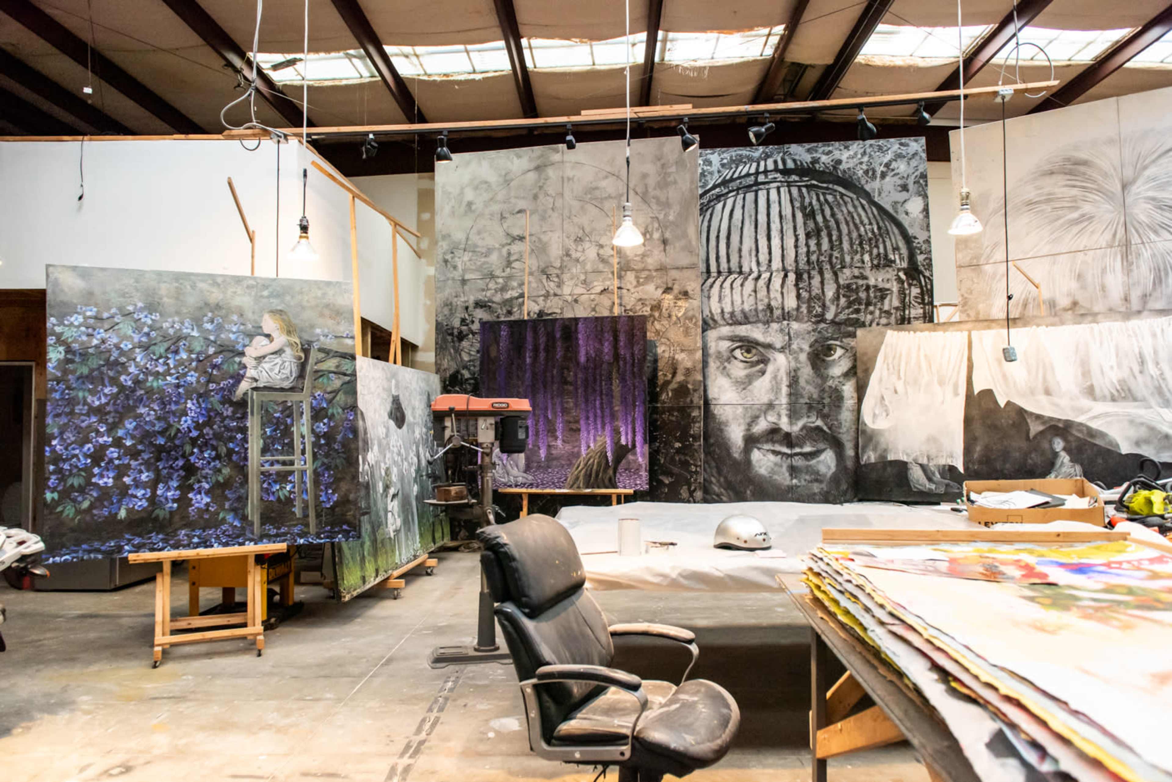 An artist's studio filled with large, unfinished canvases, a black office chair, and various art supplies scattered throughout.