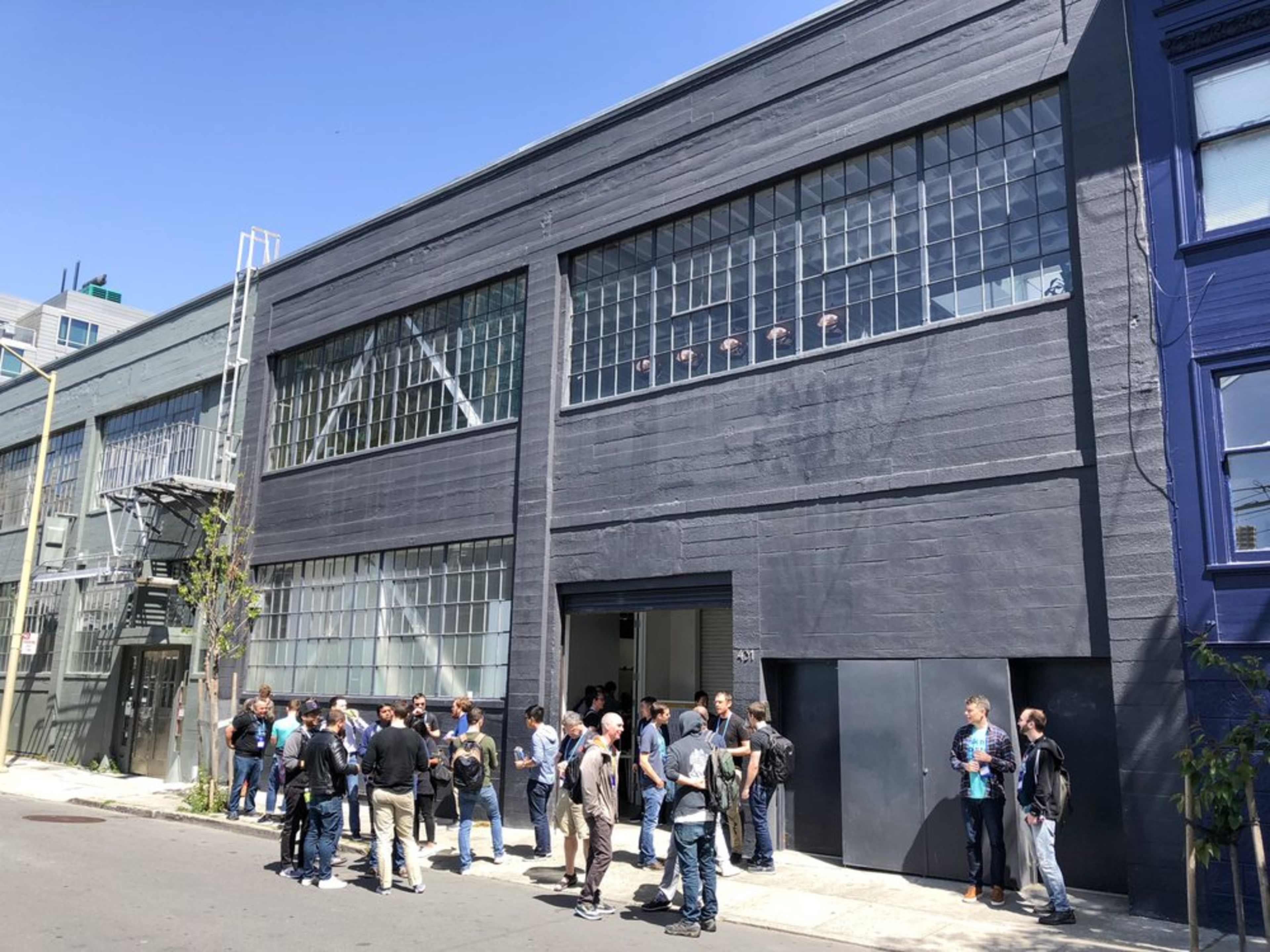 A group of people stands outside a large, gray industrial building with large windows and a double door entrance.