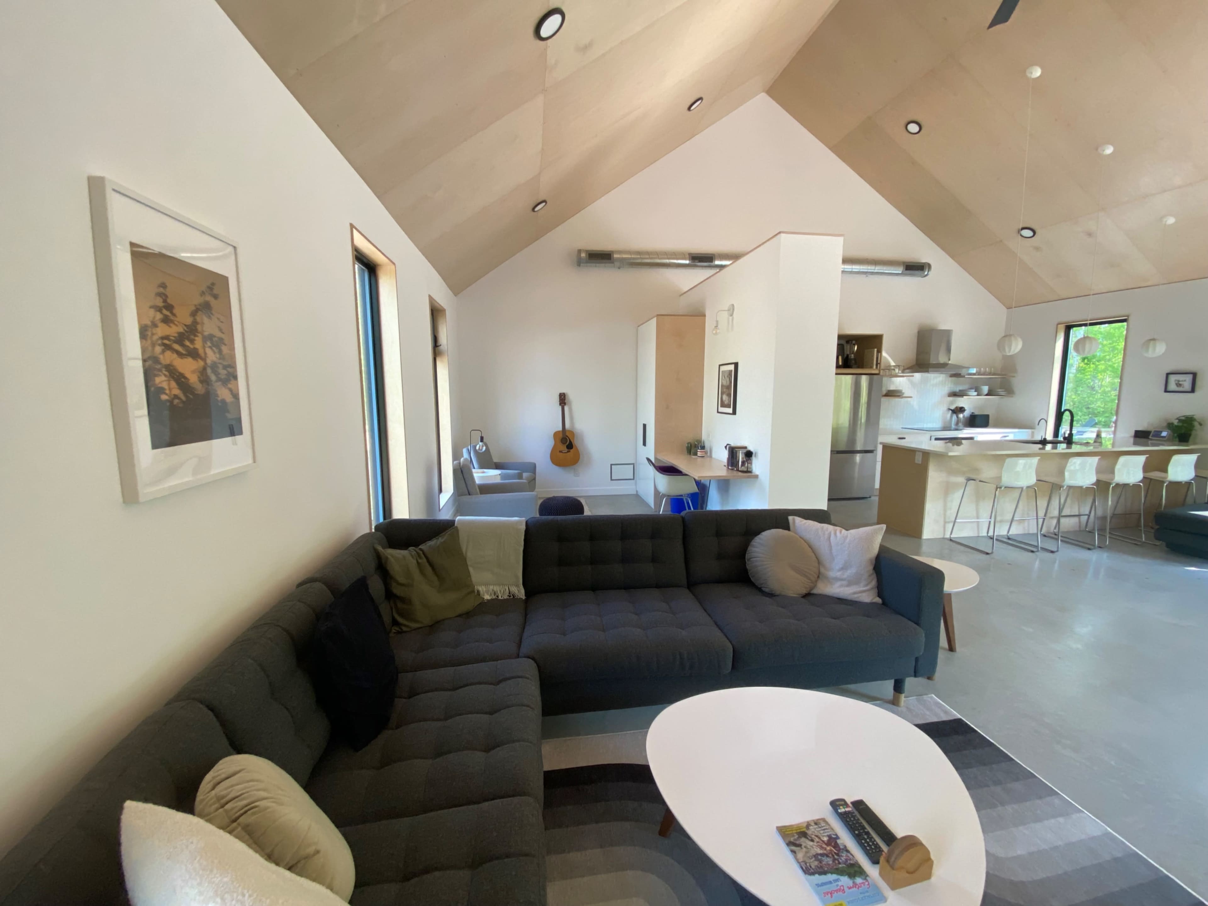 A modern living room with a sectional sofa, a coffee table, and a kitchenette in an open-concept space featuring wooden ceilings and large windows.