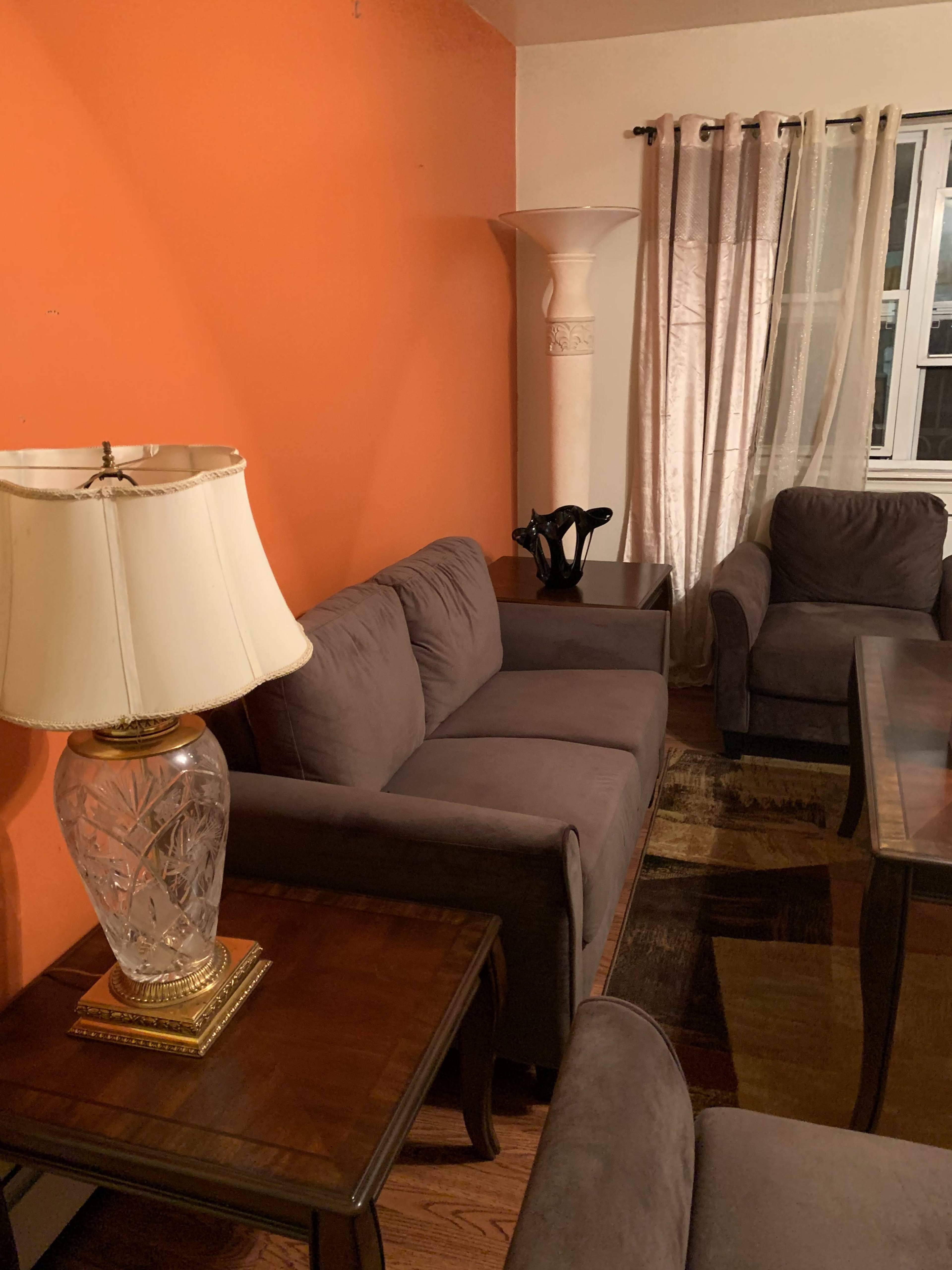 A living room with a gray sofa and armchairs, a glass lamp on a wooden table, and an orange wall with curtains next to a window.