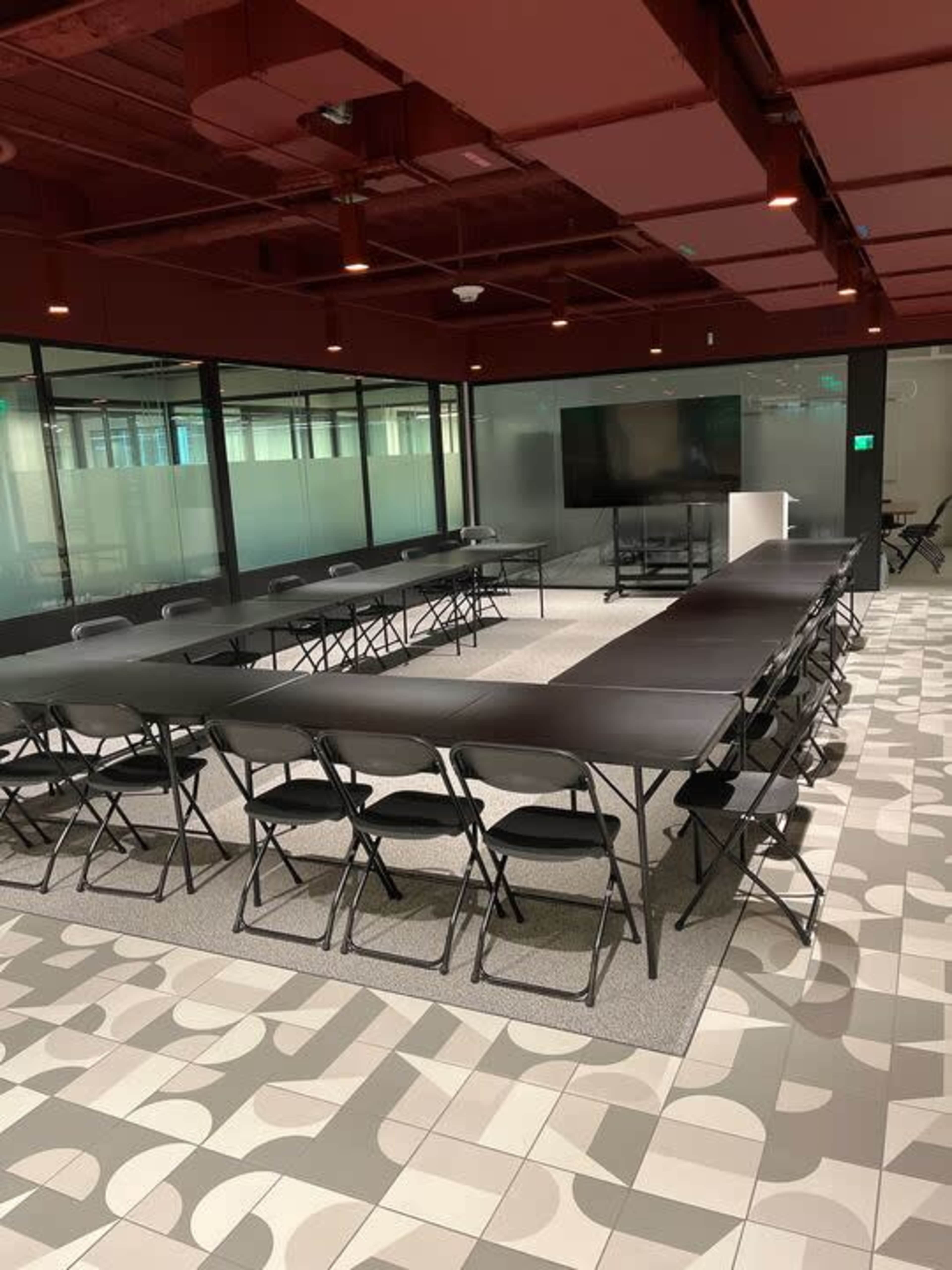 A conference room features a large rectangular table surrounded by folding chairs, with a screen and presentation area at one end.