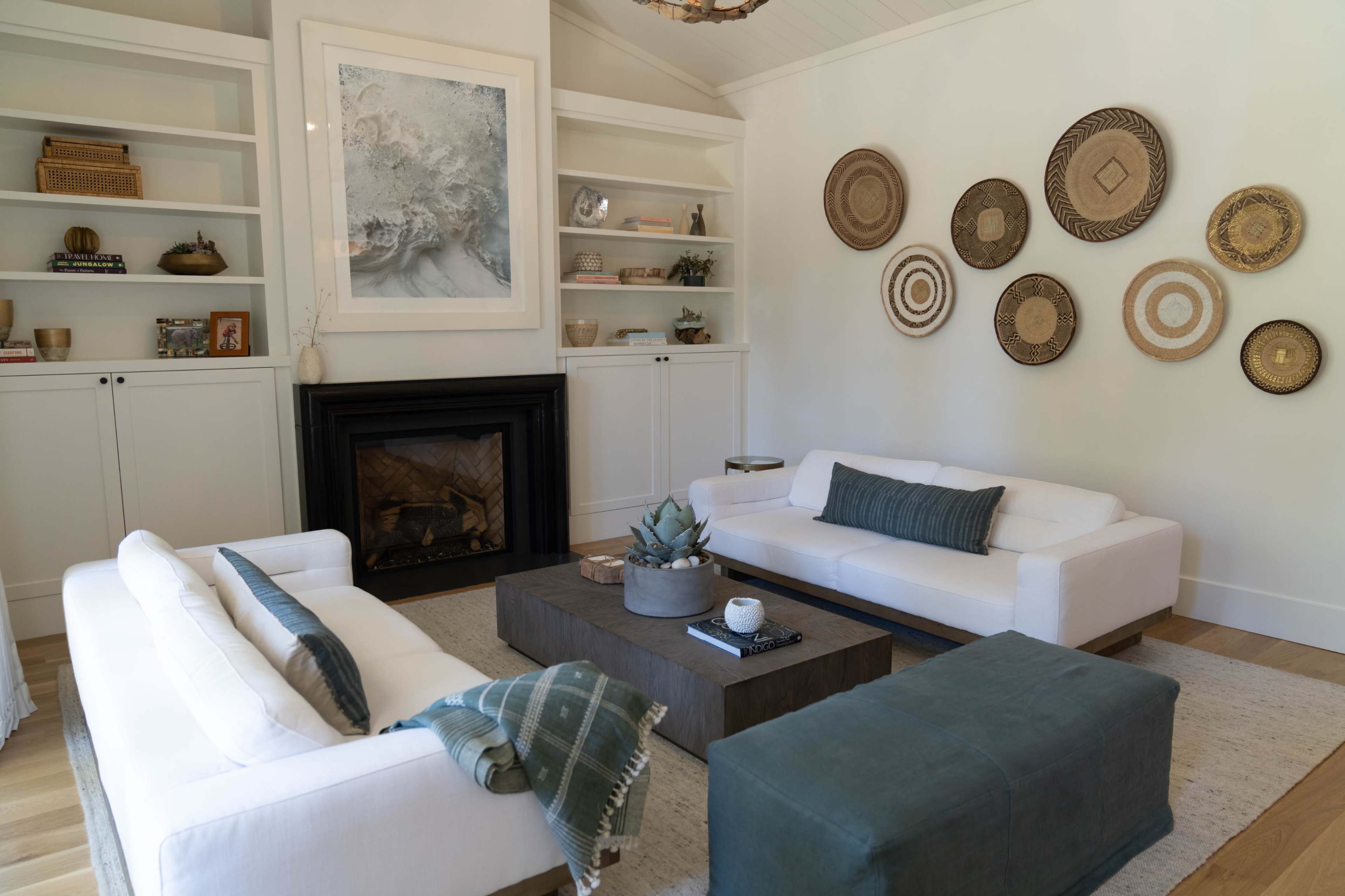 The image depicts a modern living room with two white sofas, a dark wooden coffee table, and woven wall decor.