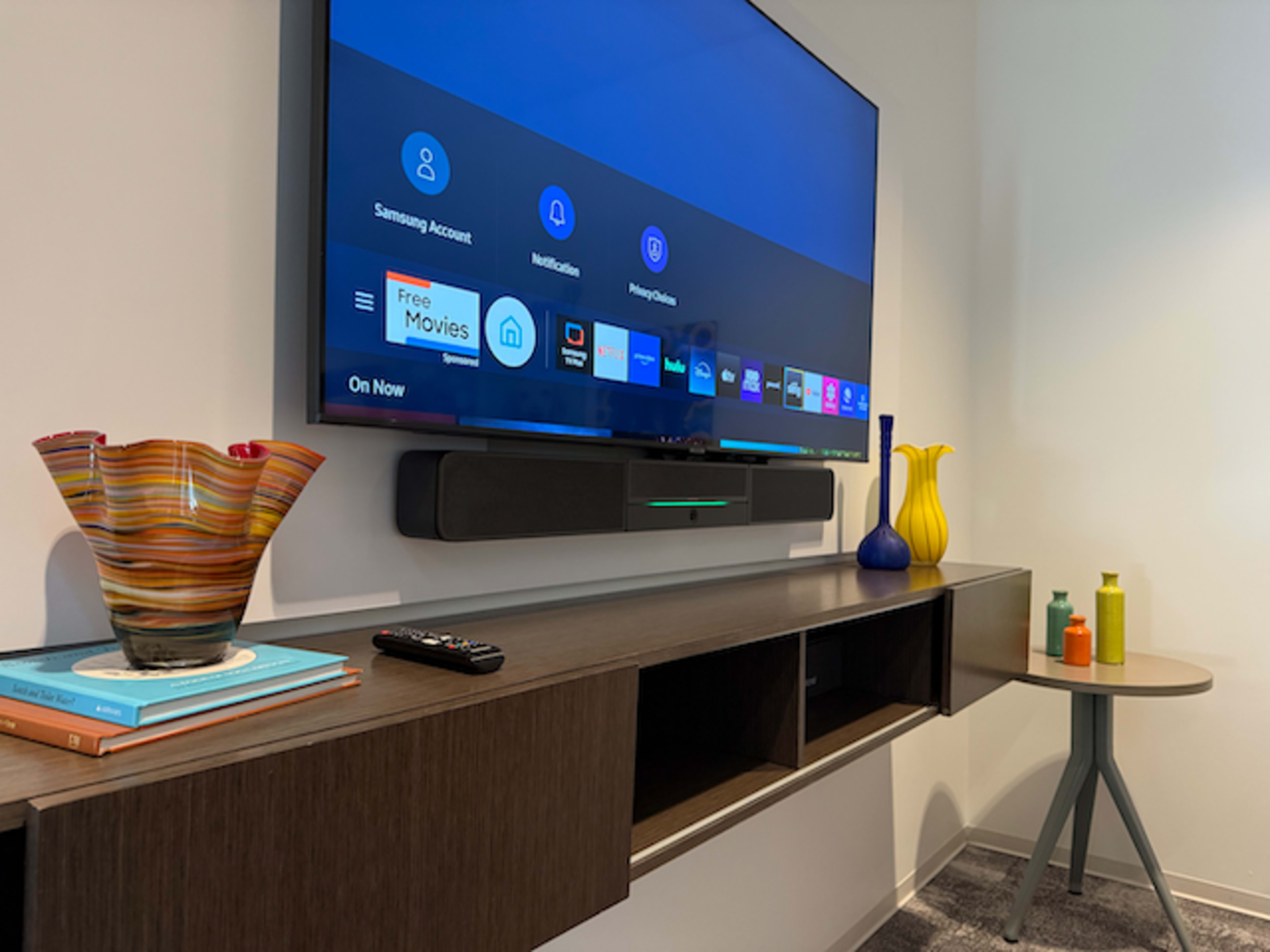 A sleek mounted television displays a user interface on a light-colored wall above a modern wooden entertainment unit with decorative vases and books.