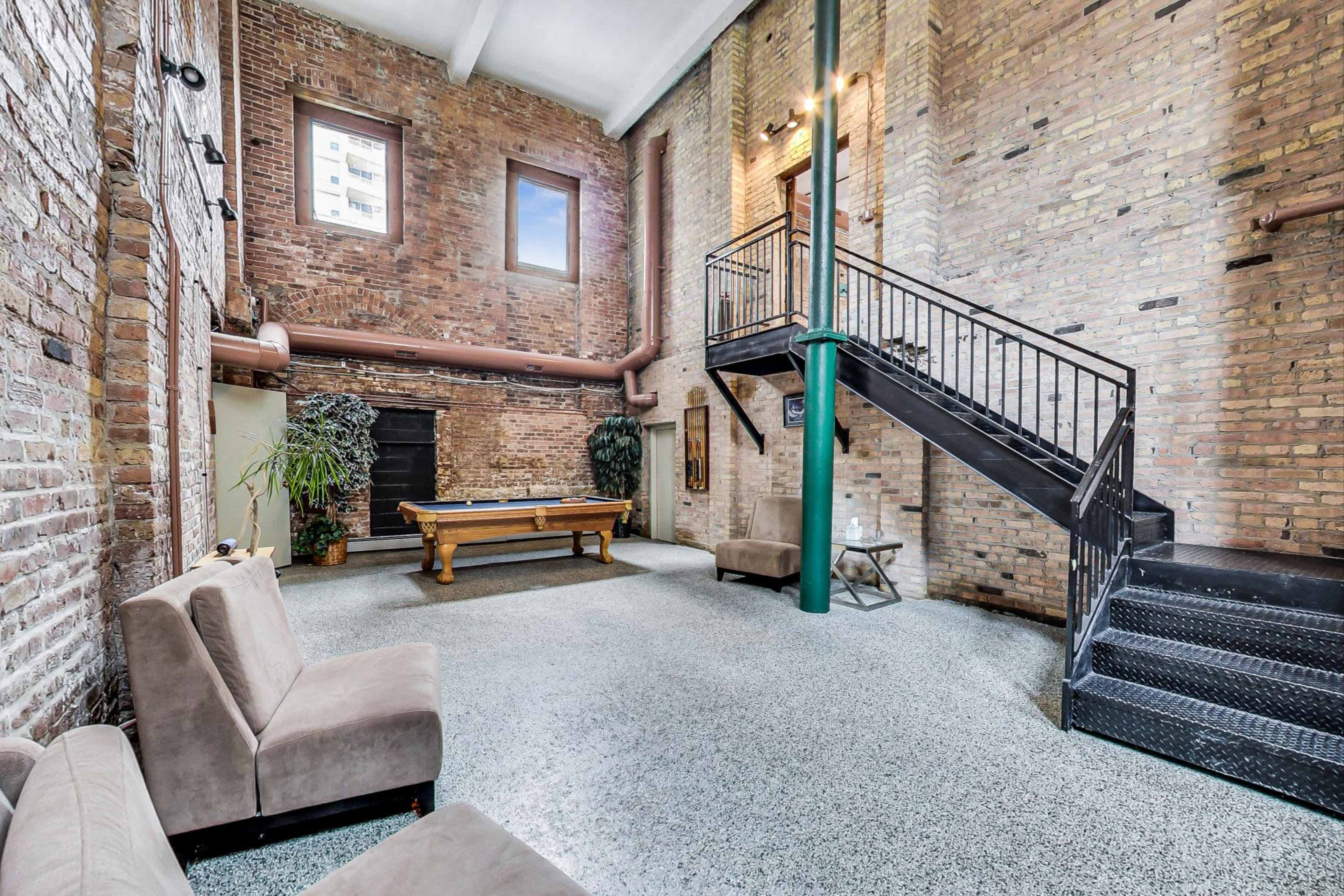 An industrial-style room featuring exposed brick walls, a staircase, and a pool table, with seating arranged to the side.