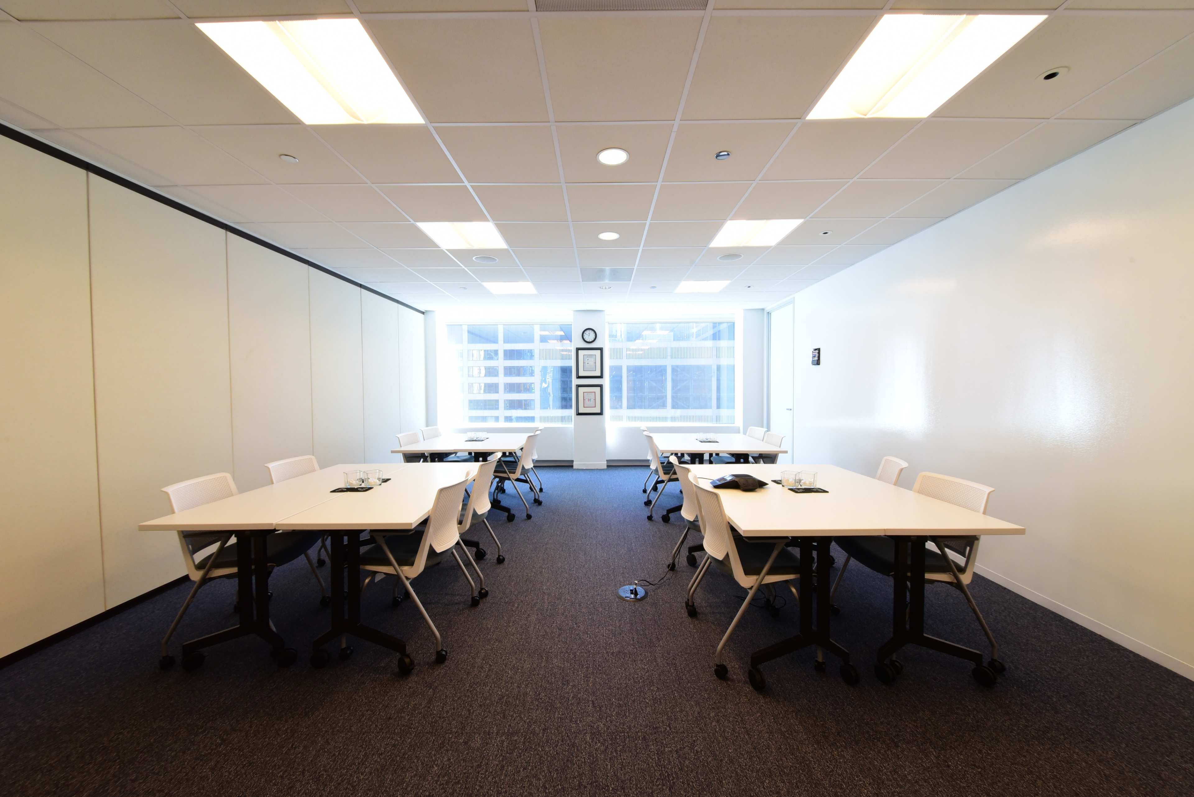 A conference room features several white tables arranged in a symmetrical layout, with chairs positioned around them and large windows allowing natural light.