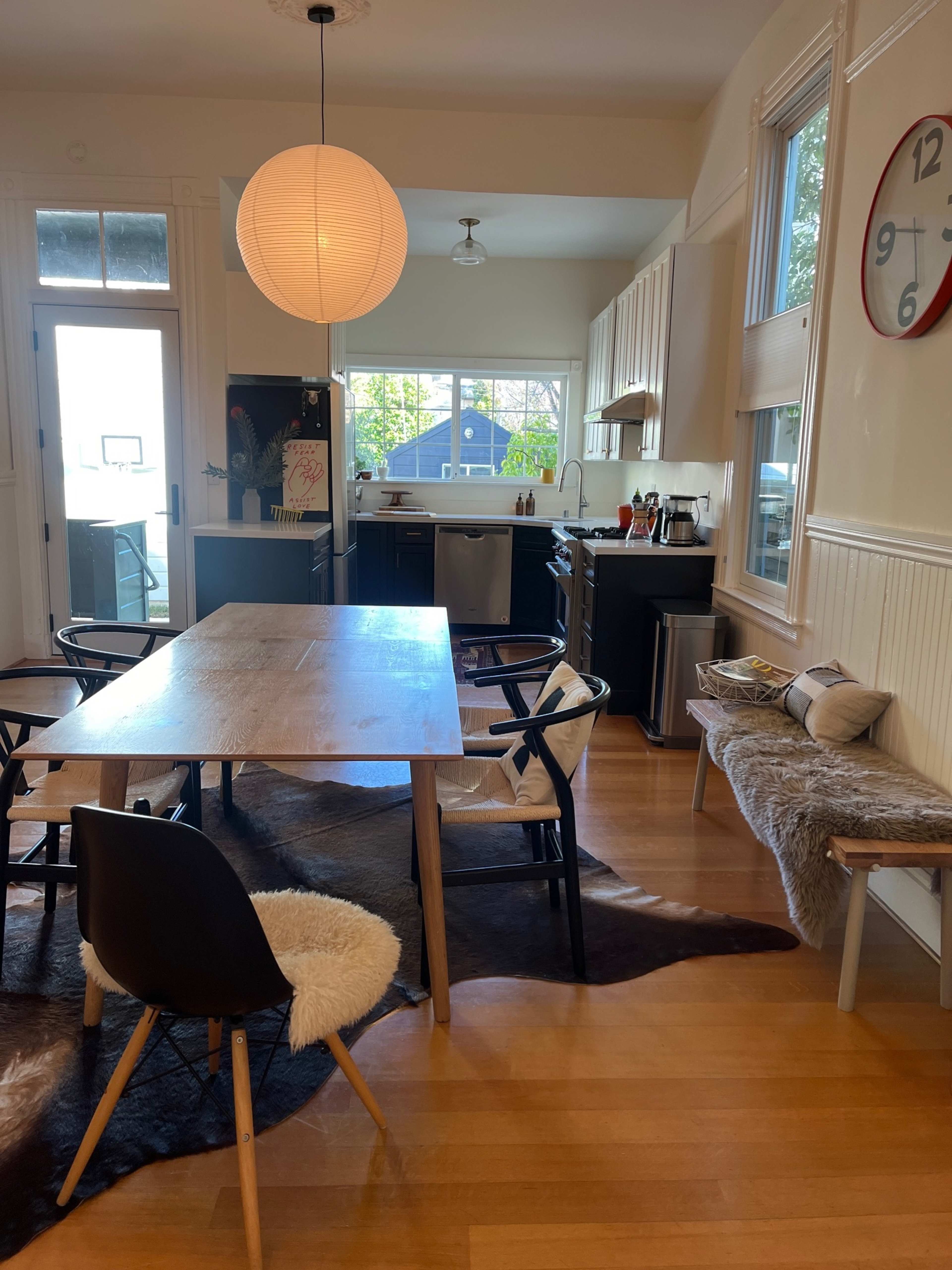 A bright kitchen and dining area featuring a wooden table, black and white chairs, and a large round lamp hanging from the ceiling.