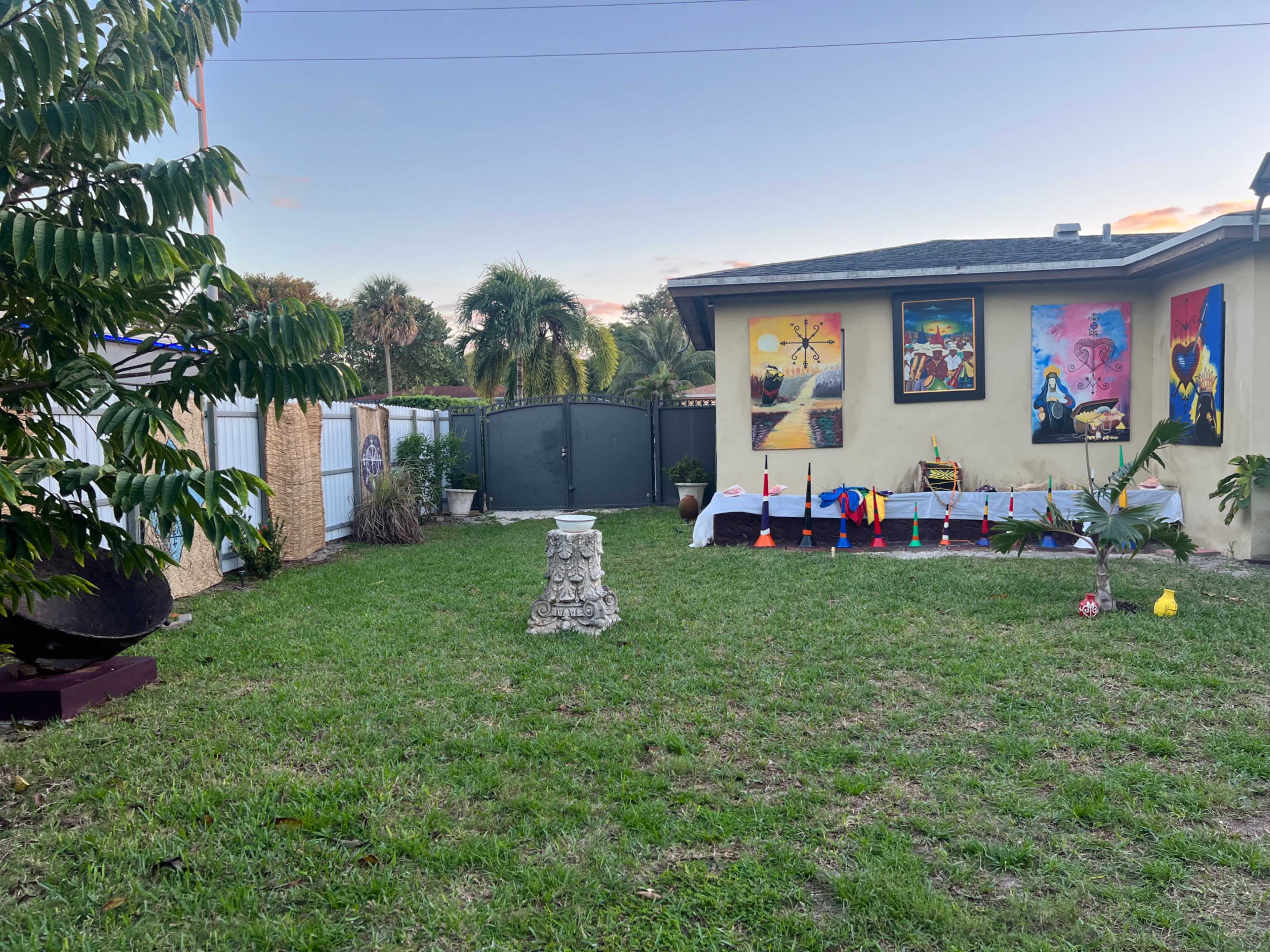 A backyard with a grassy area featuring a stone pedestal, colorful decorations, and a wall adorned with several framed artworks.