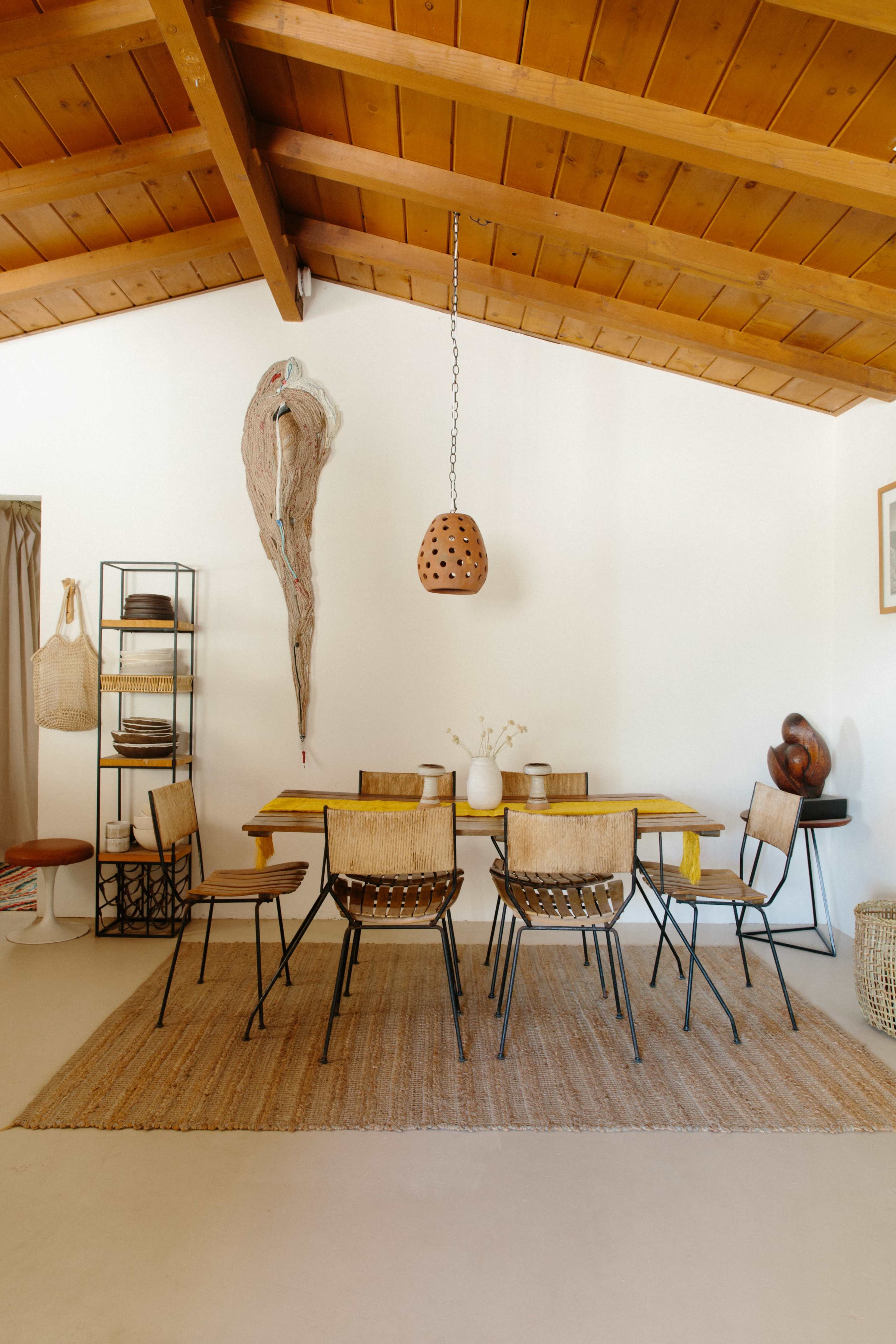 A dining area features a large, wooden table surrounded by chairs, with a suspended light fixture and decorative wall elements in a room with a vaulted ceiling.