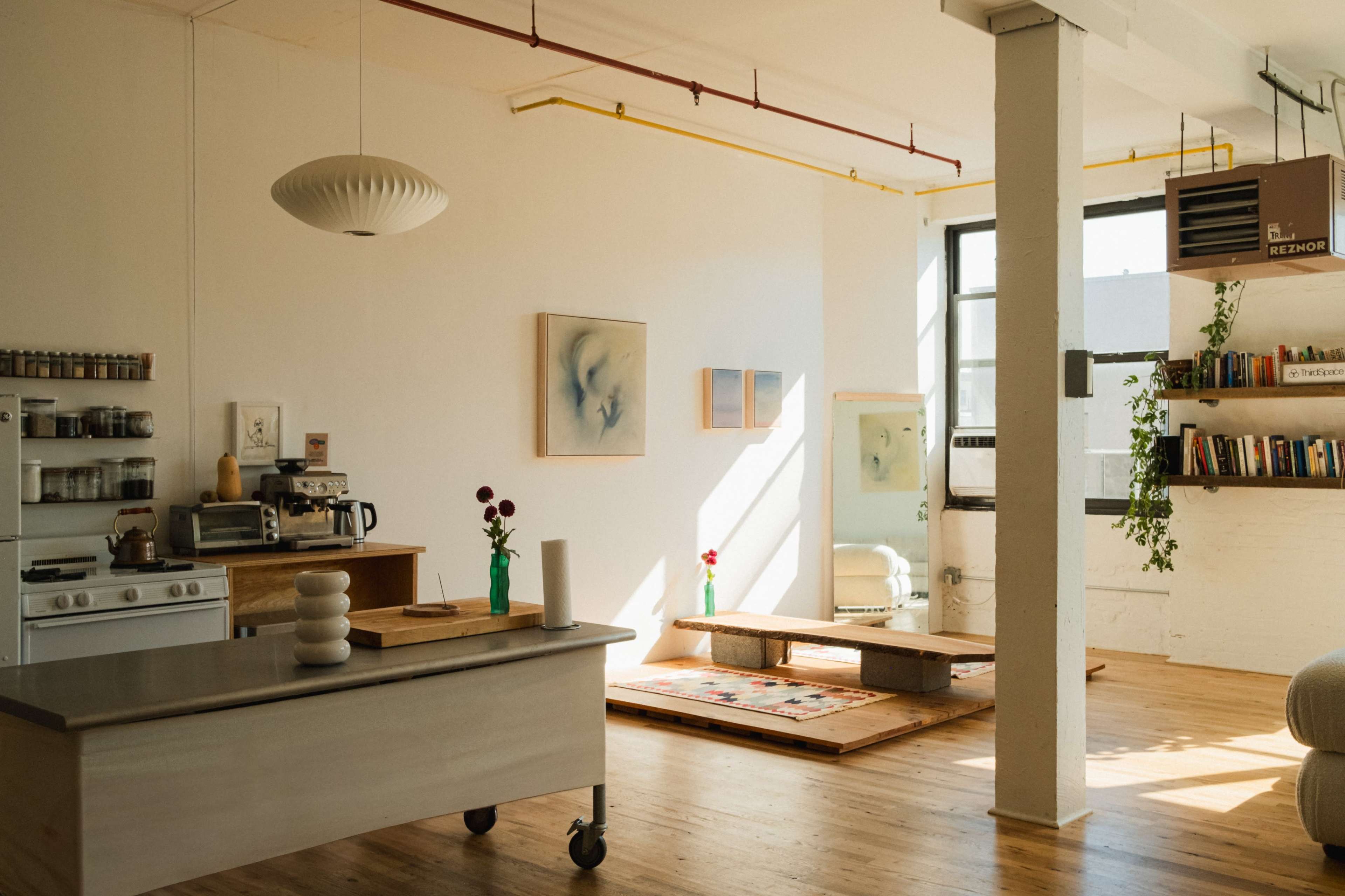 The image shows a bright, minimalist interior of a modern living space featuring a kitchen area, wooden floors, and a small table with a vase of flowers.