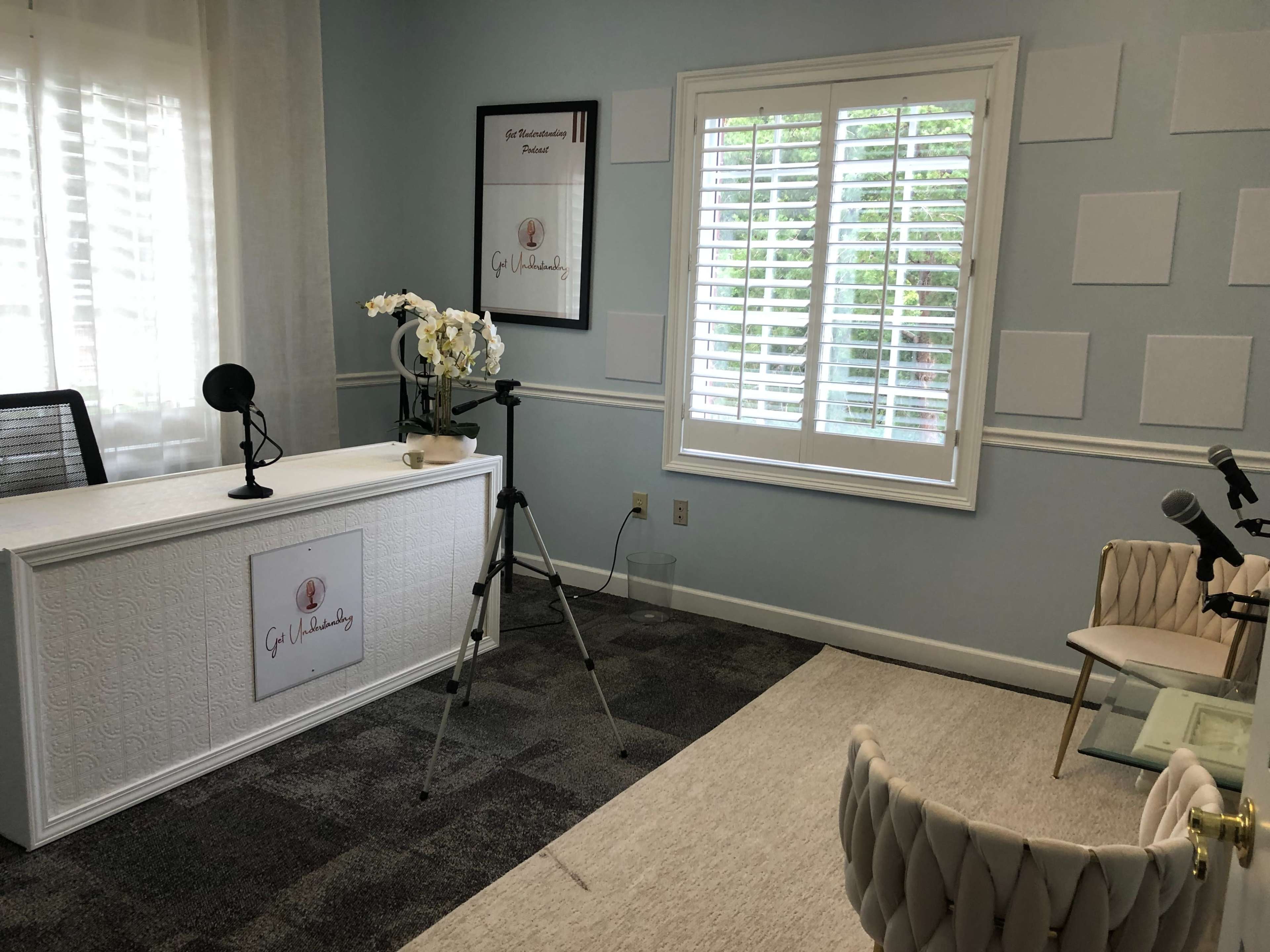 The image shows a light blue room with a desk featuring a logo, a microphone setup, a camera on a tripod, and a white chair.