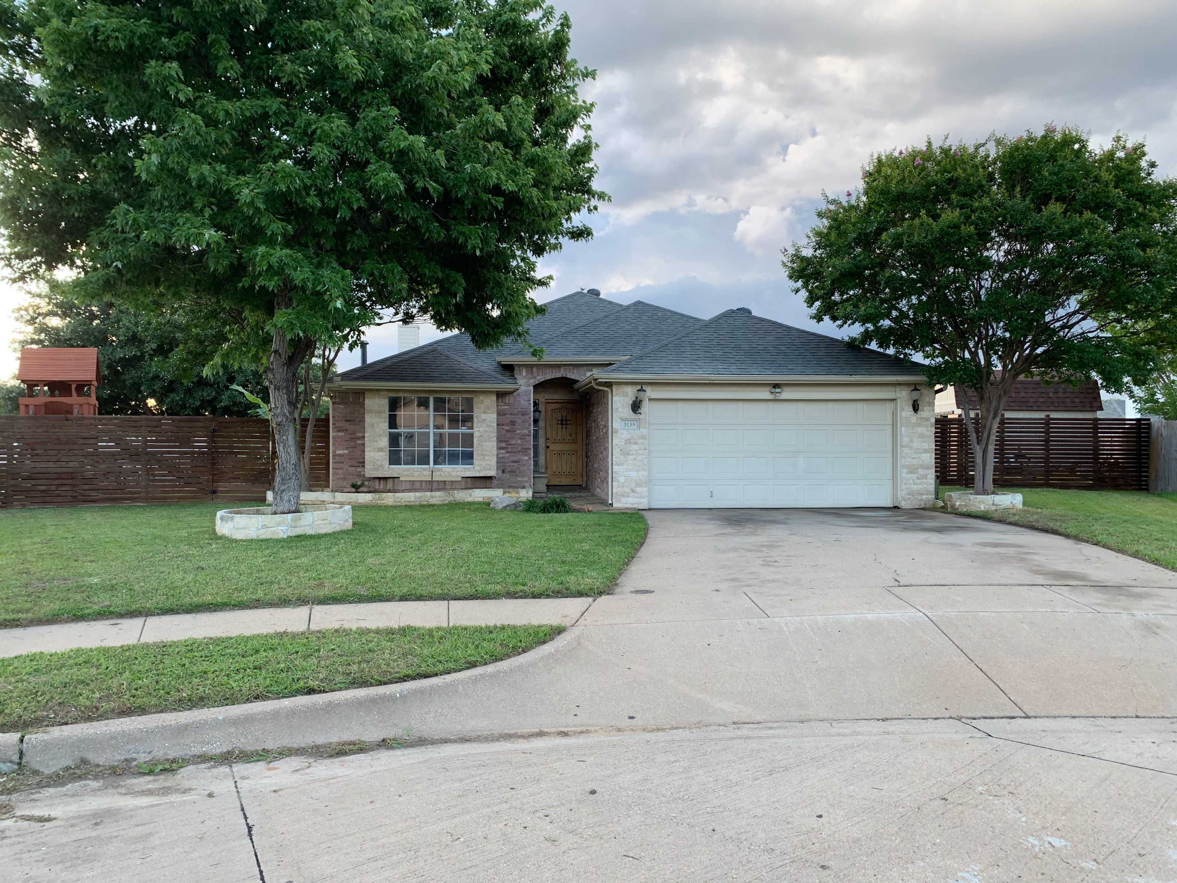 A single-story house with a stone facade and a two-car garage is situated on a curved driveway, surrounded by grassy landscaping and trees.