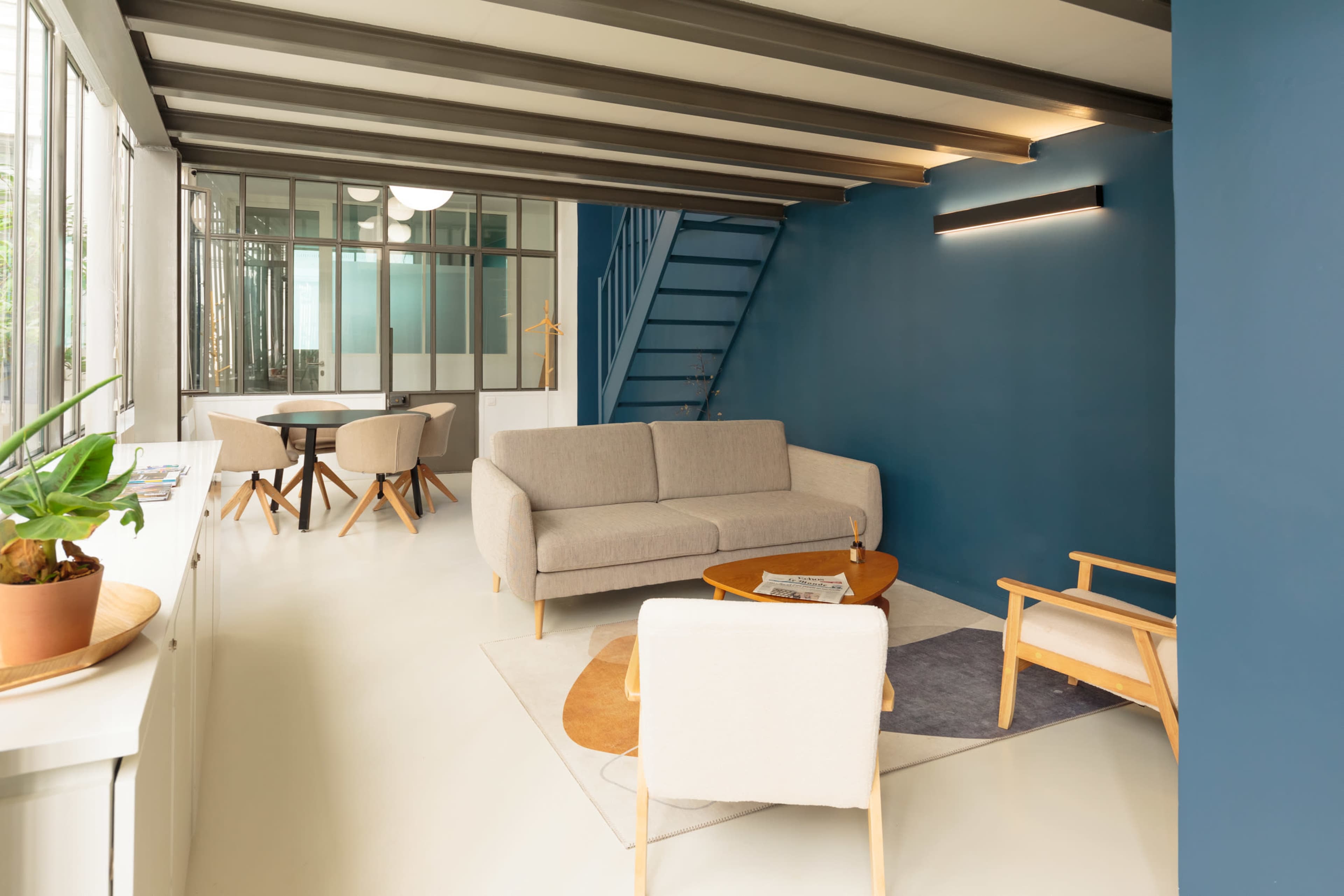 The image shows a modern living space with a gray sofa, light chairs, a wooden table, and a dark blue accent wall, all illuminated by natural light from large windows.