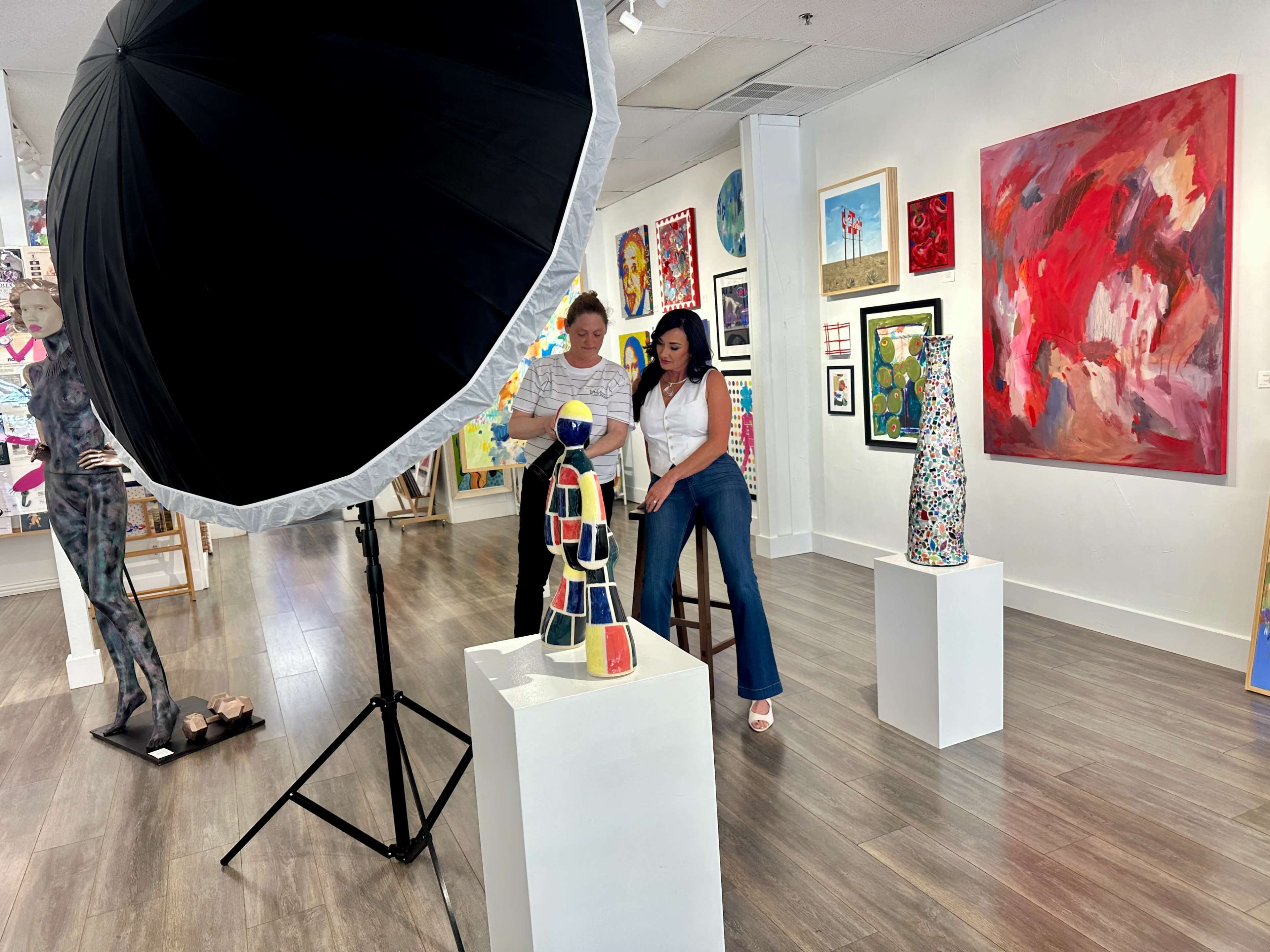 Two individuals pose for a photo in an art gallery, surrounded by various colorful artworks and sculptures.