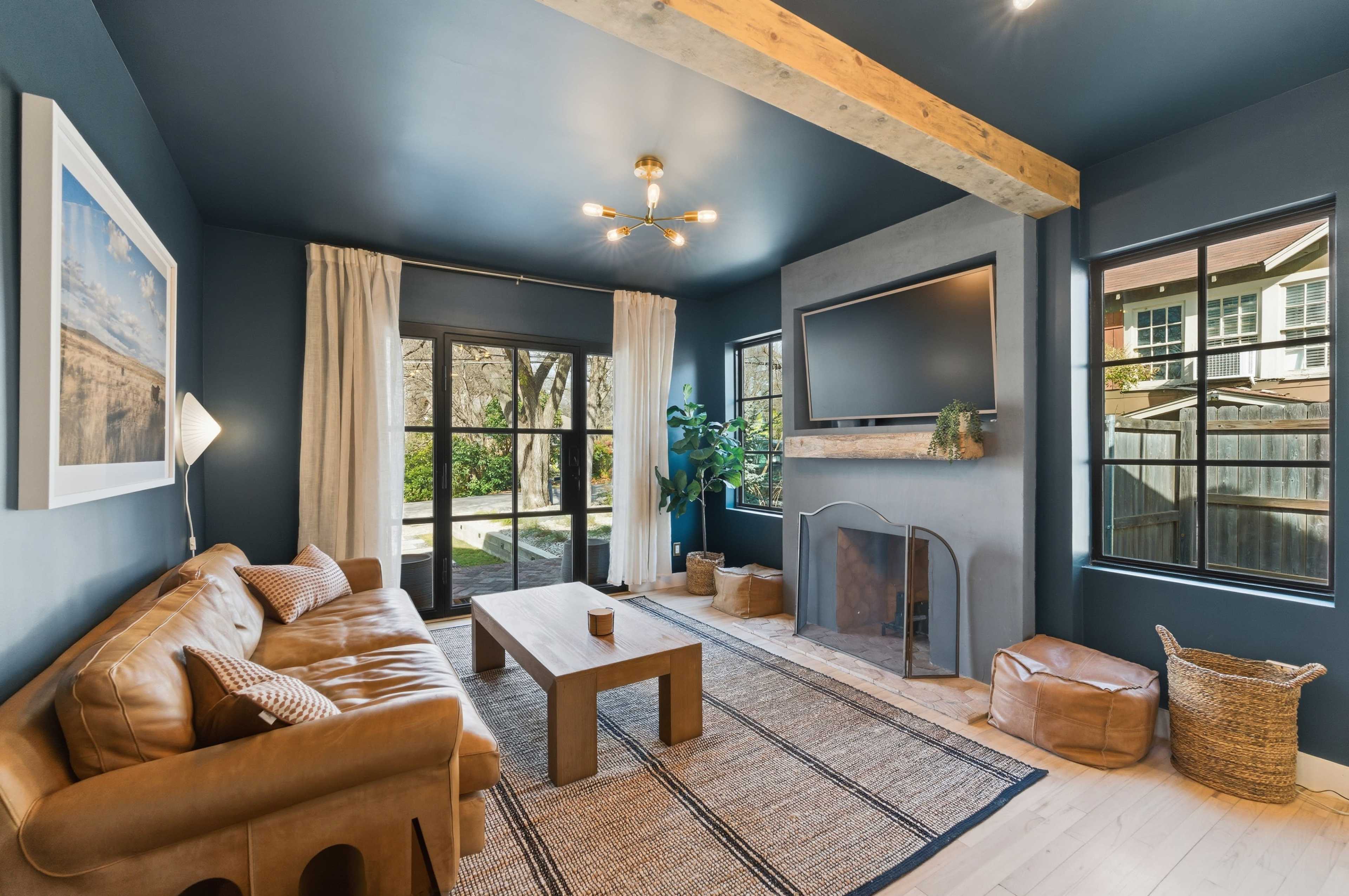 The image shows a modern living room featuring a brown leather sofa, a wooden coffee table, and a rug on light-colored flooring, with large windows allowing natural light to illuminate the space.