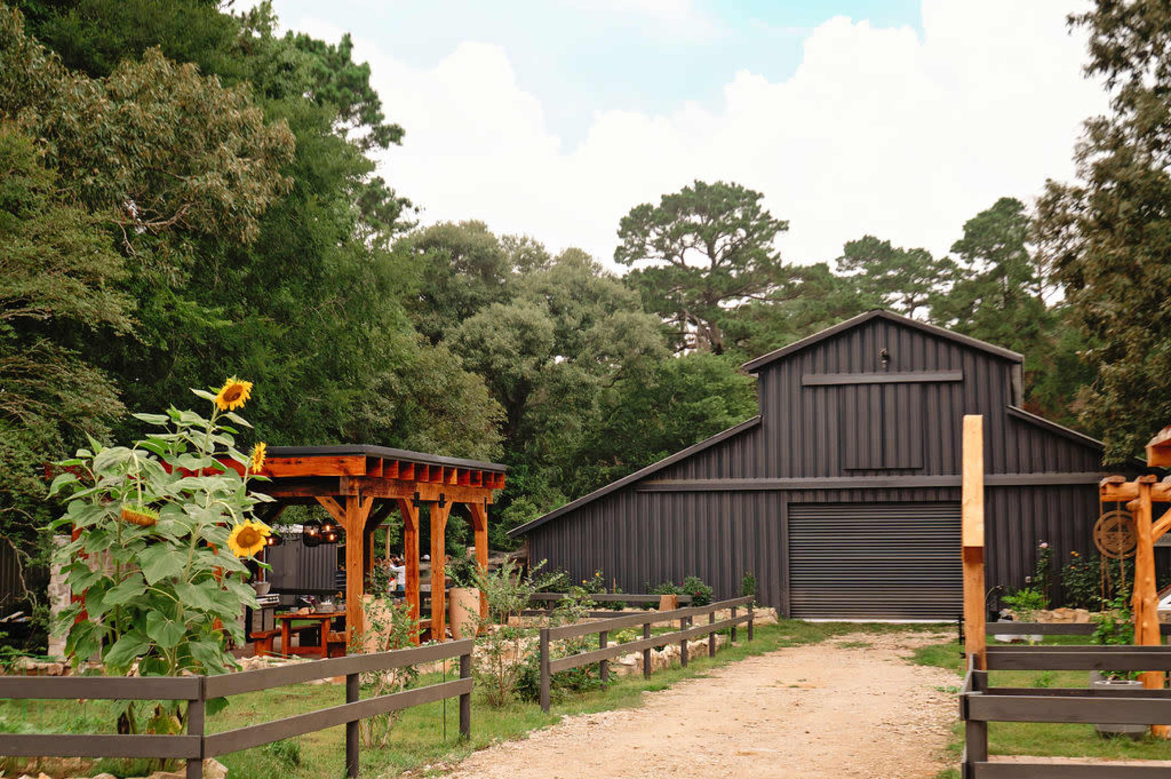 A gravel driveway leads to a large, dark-colored barn surrounded by trees and sunflowers.