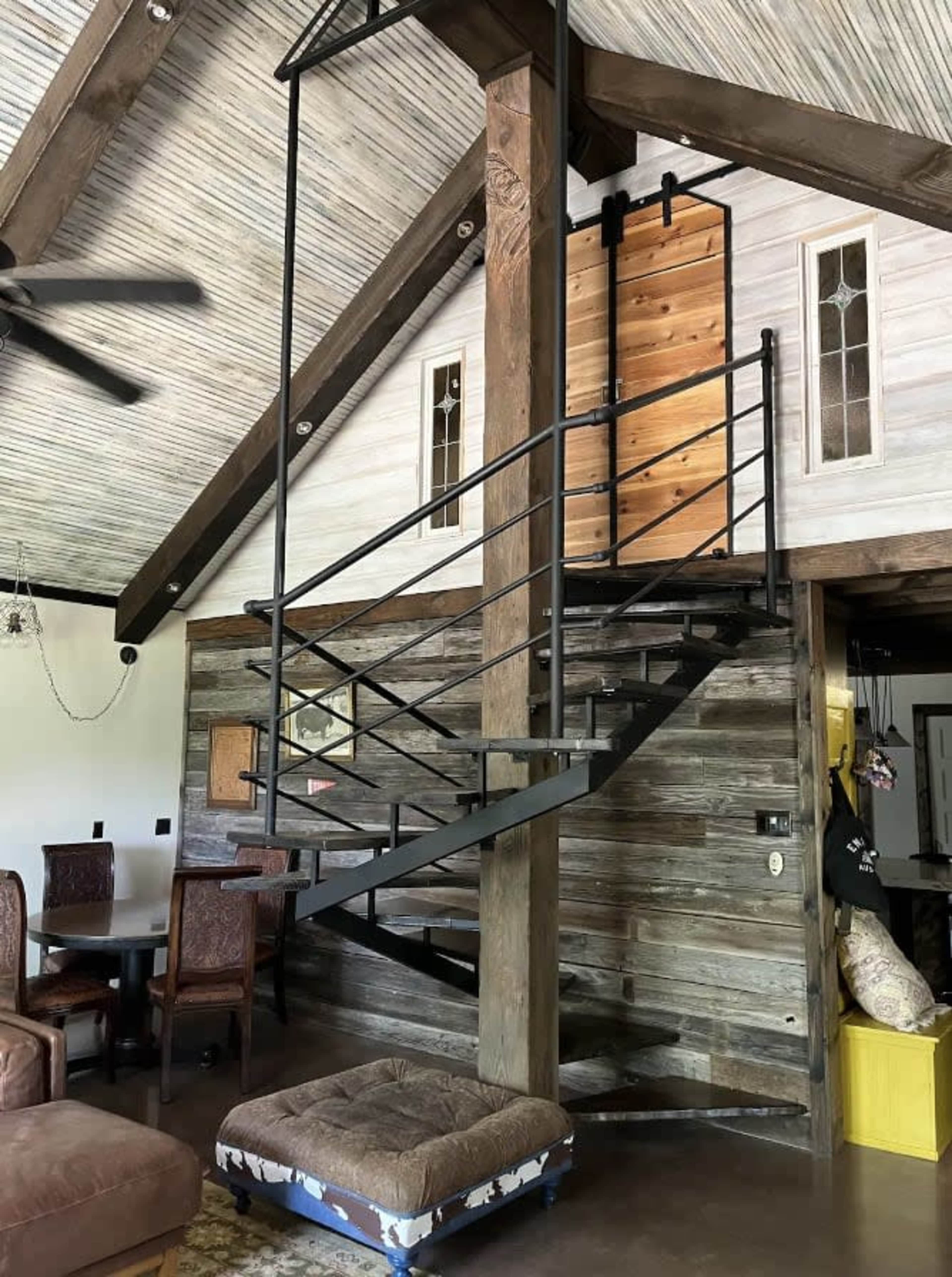 The image shows a spiral staircase made of metal, leading to an upper level in a rustic-style home with wooden walls and a partially open design.