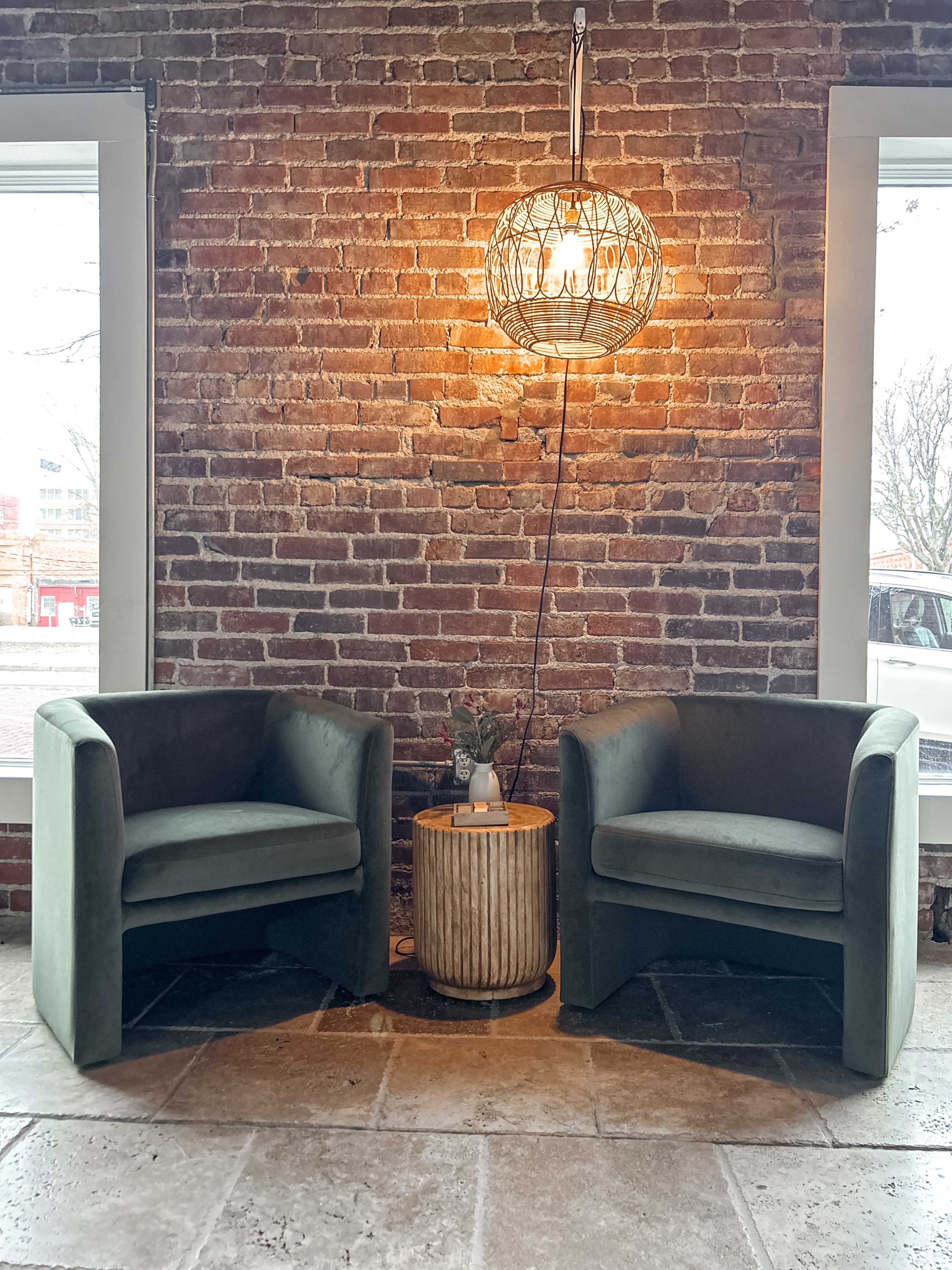 Two green velvet chairs are positioned beside a round wooden side table under a metal pendant light, against a backdrop of exposed brick walls and windows.