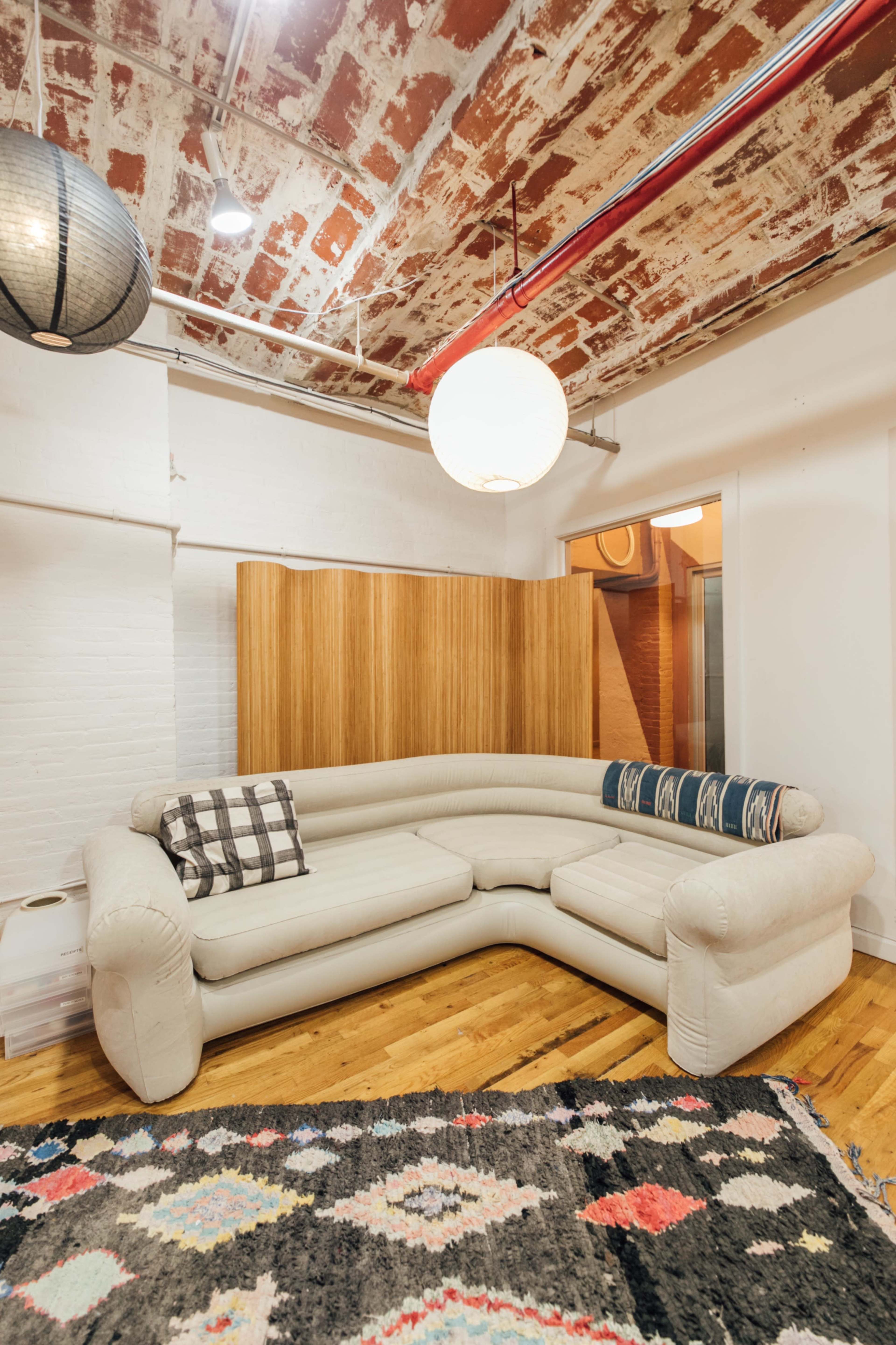 The image shows a modern, light-colored sectional sofa positioned on a patterned rug in a room with exposed brick walls and overhead lighting.