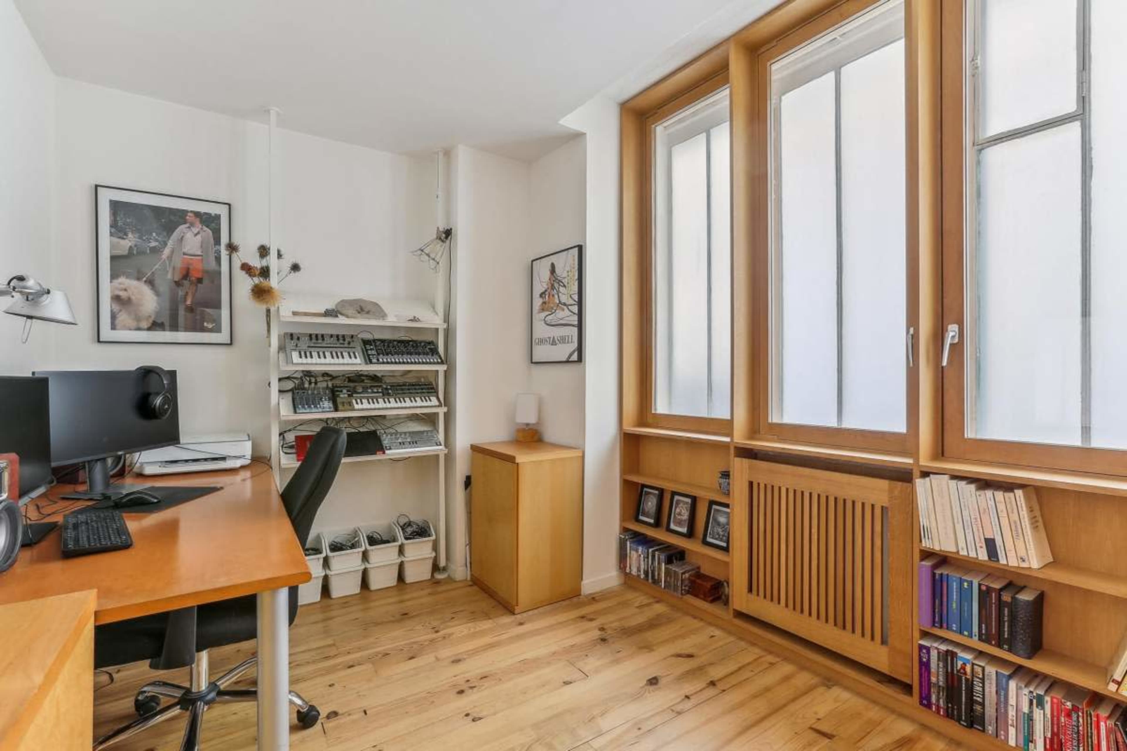 The image shows a bright home office featuring a wooden desk with a computer, shelves holding musical equipment, and large windows allowing natural light to enter the space.