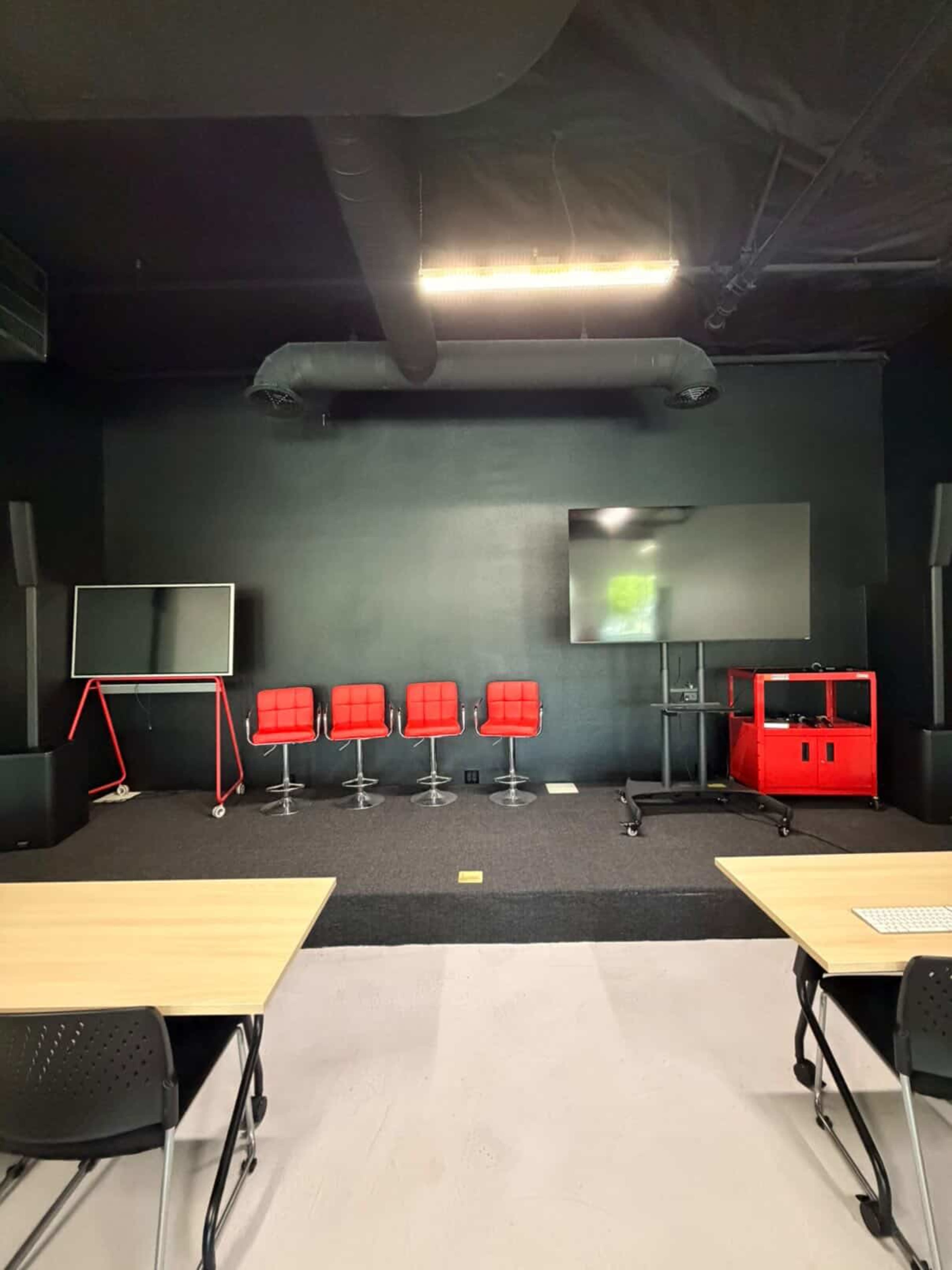 The image shows a modern meeting room with two large screens, four red chairs on a black floor, and two wooden tables in front.