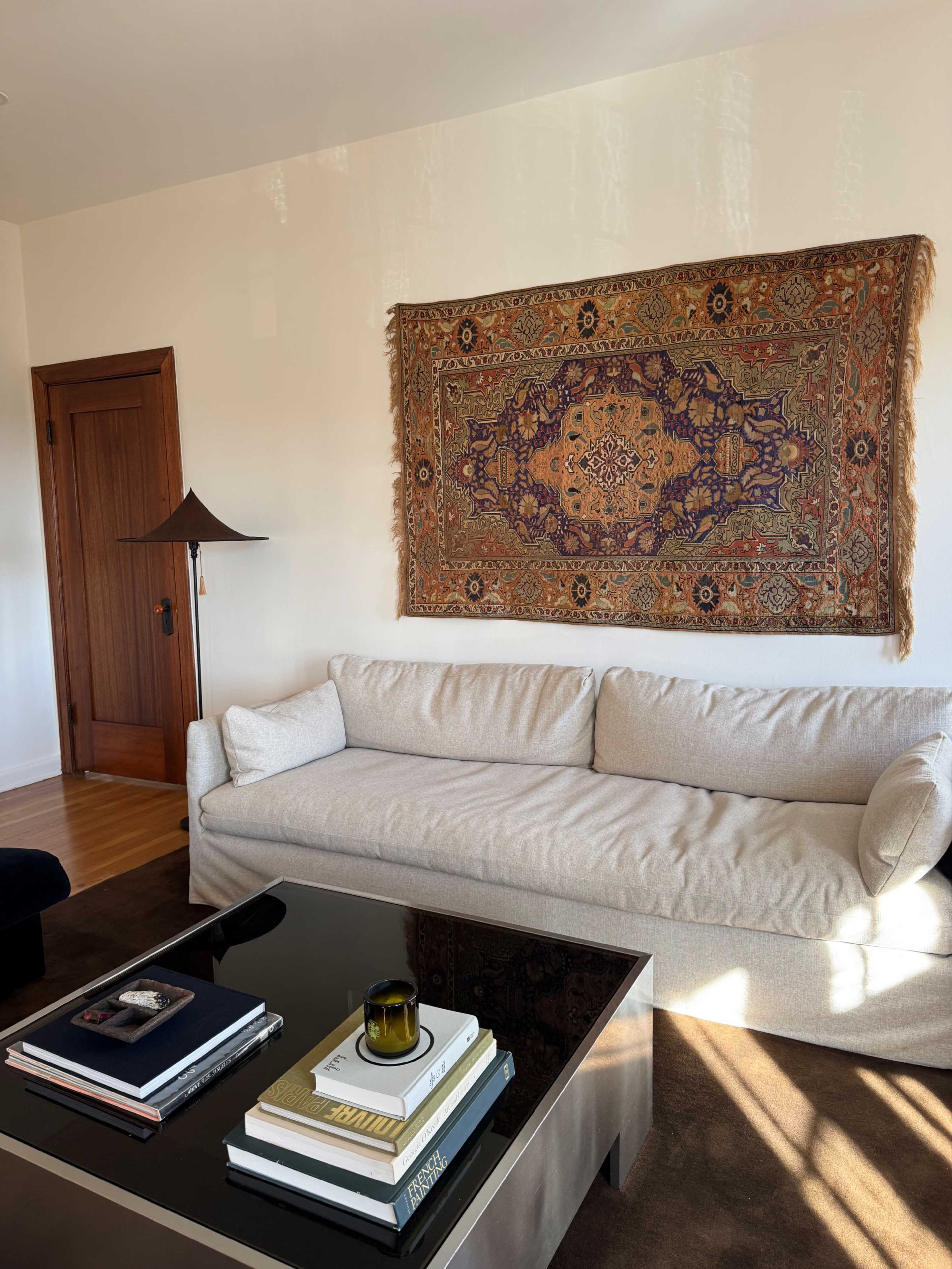 A beige sofa sits in a sunlit room with a large decorative rug hanging on the wall and a glass coffee table stacked with books and a candle.