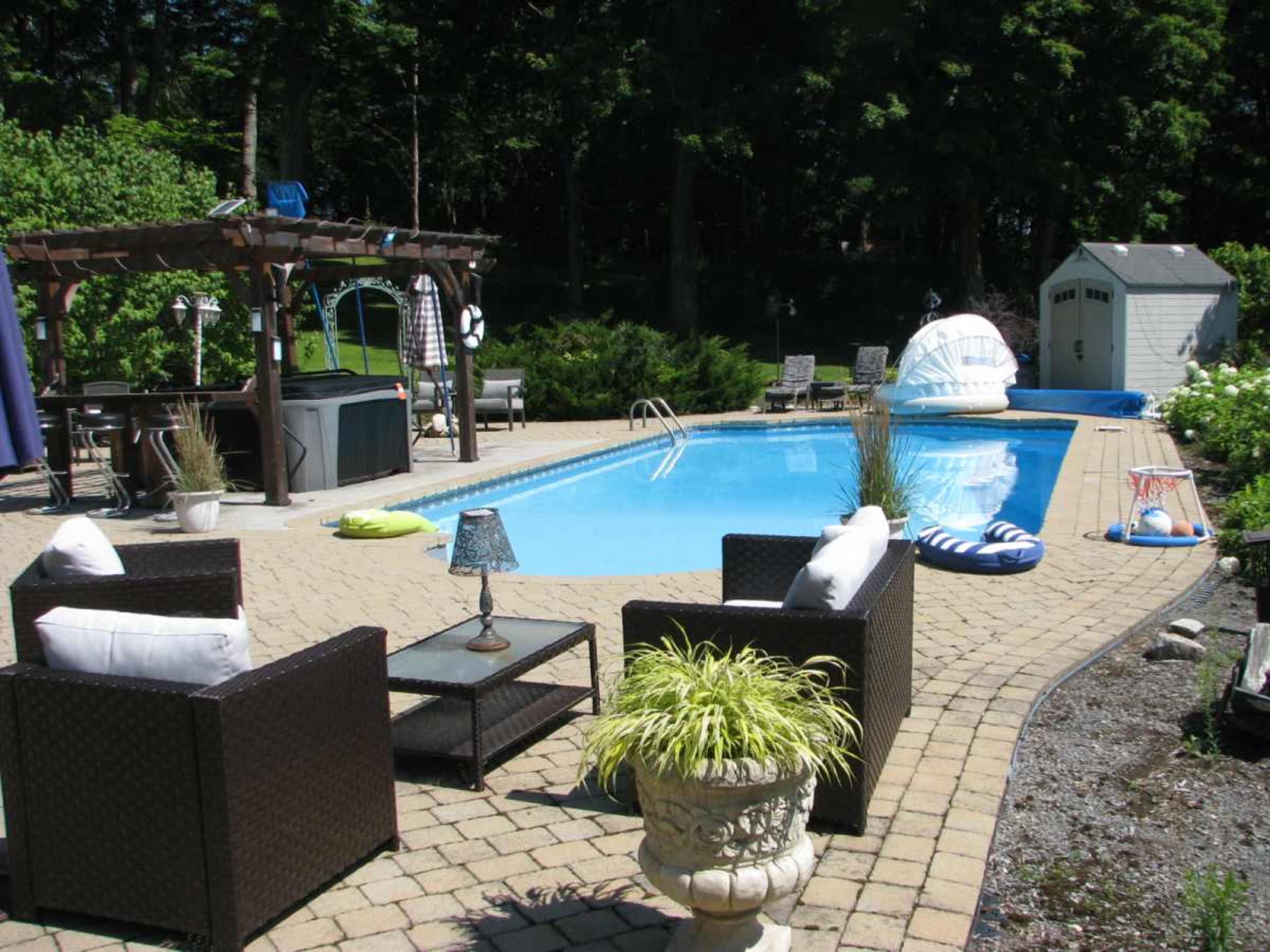 A backyard scene featuring a rectangular swimming pool surrounded by decorative landscaping, seating areas with wicker furniture, and a small shed in the background.