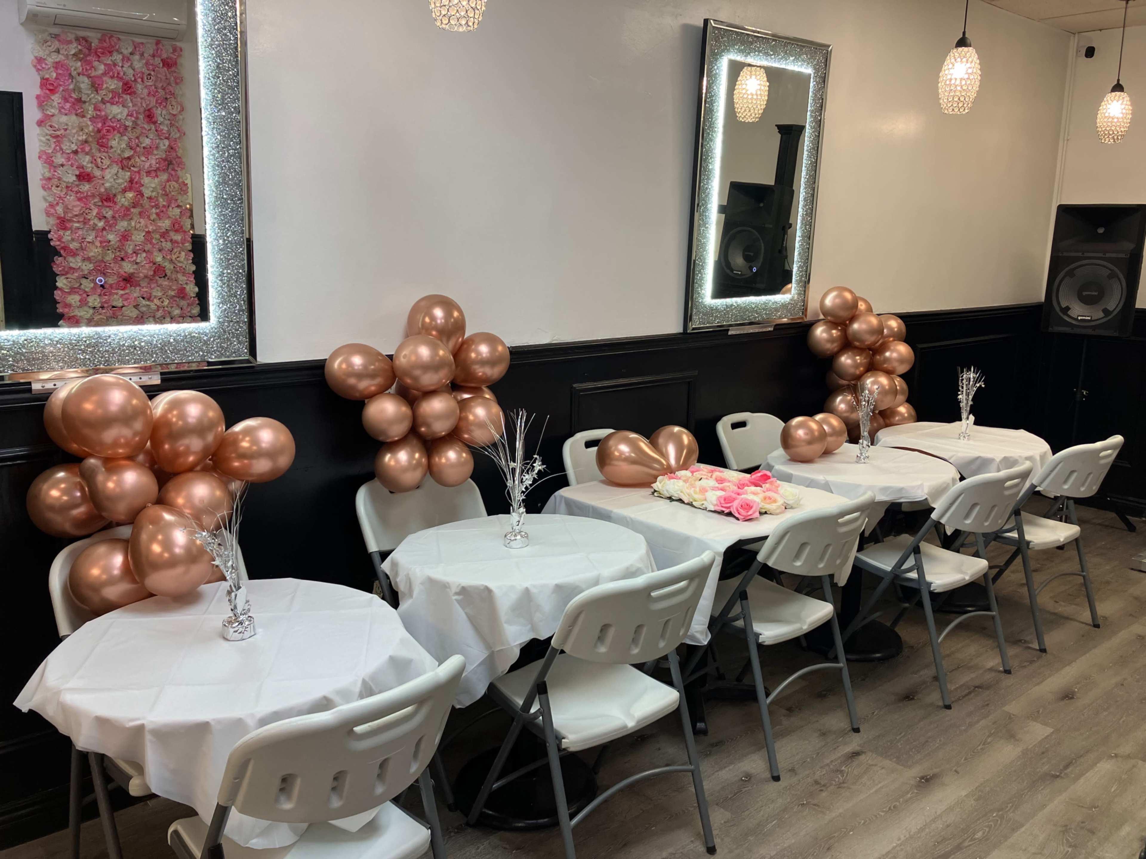 The image shows a decorated indoor event space with tables covered in white cloth, adorned with rose gold balloons and floral arrangements.