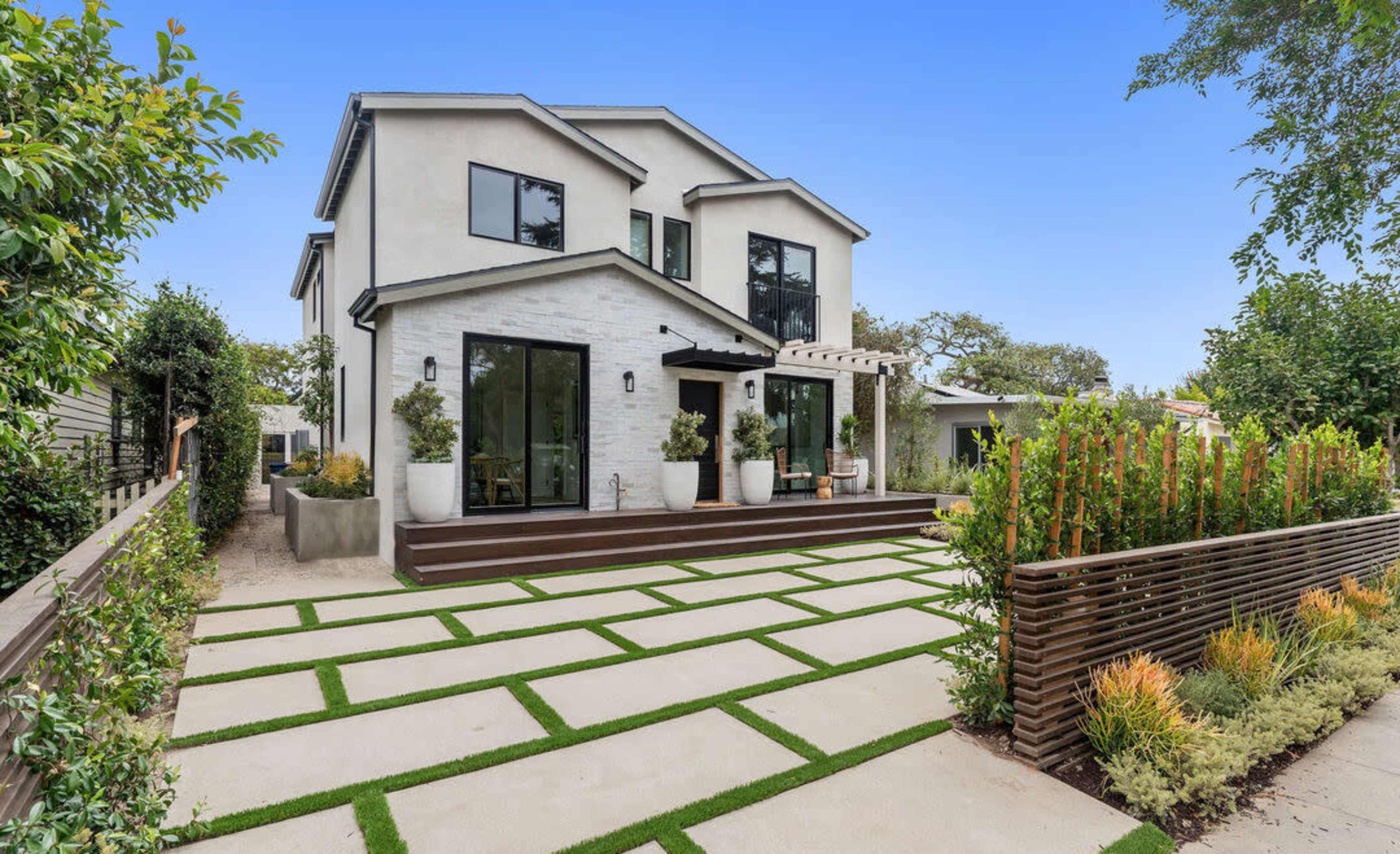 The image shows a two-story modern house with large windows, a patio area, and a landscaped yard featuring geometric pathways and plants.