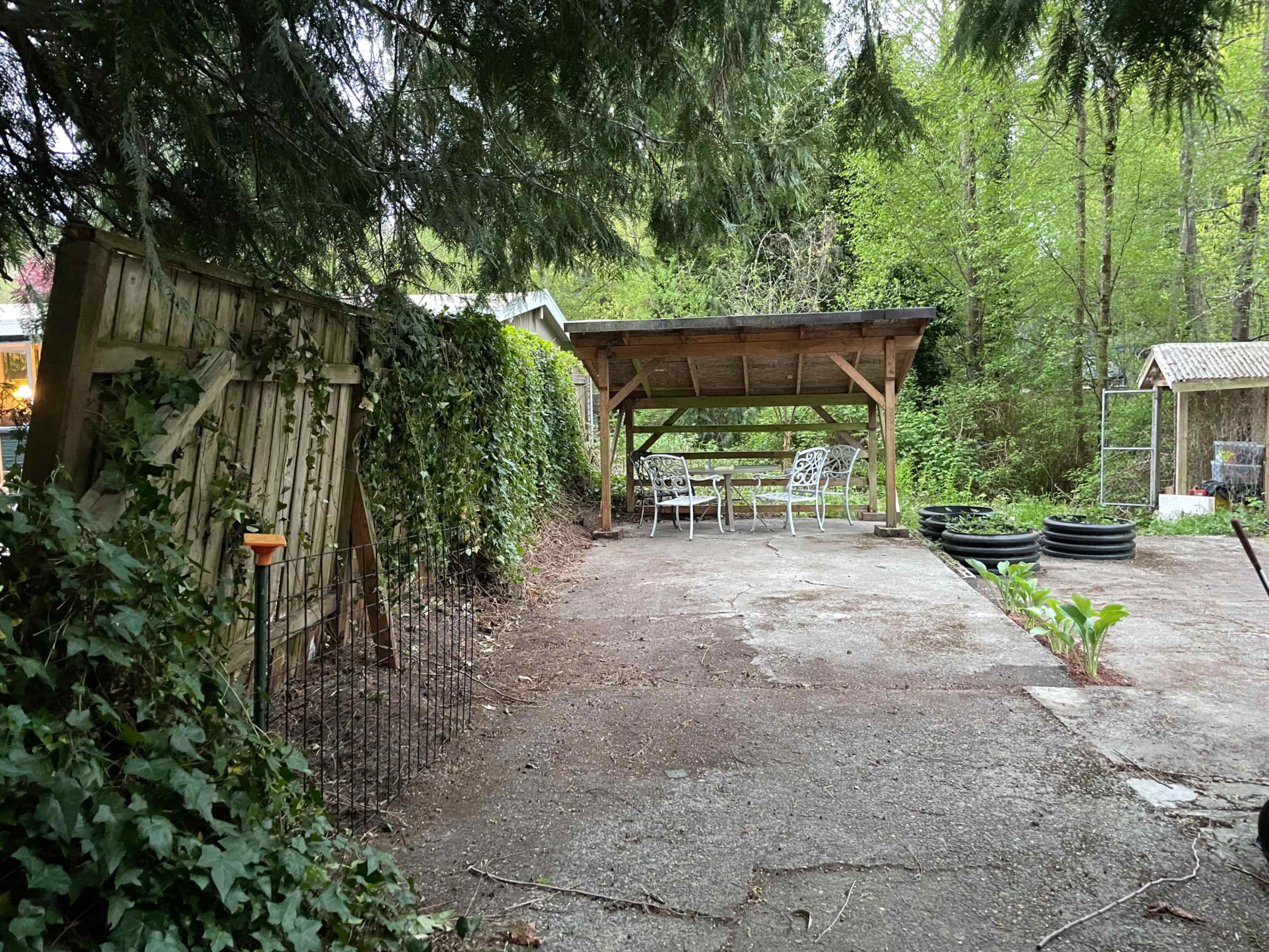 A gravel area in a wooded garden featuring a covered seating space with white metal chairs and tables, surrounded by greenery and fencing.
