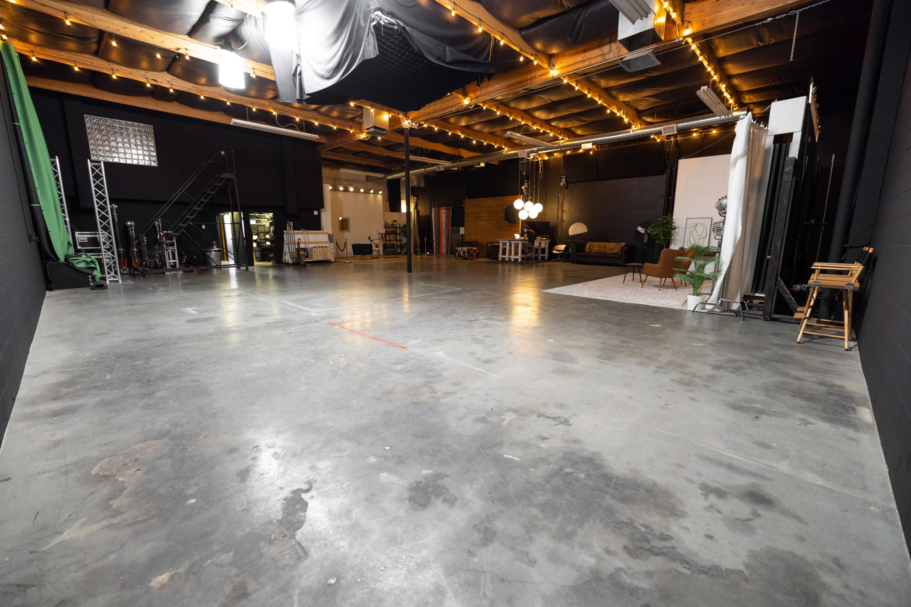 The image shows a large, empty studio space with concrete flooring, exposed ceiling beams, and scattered furniture along the back wall.